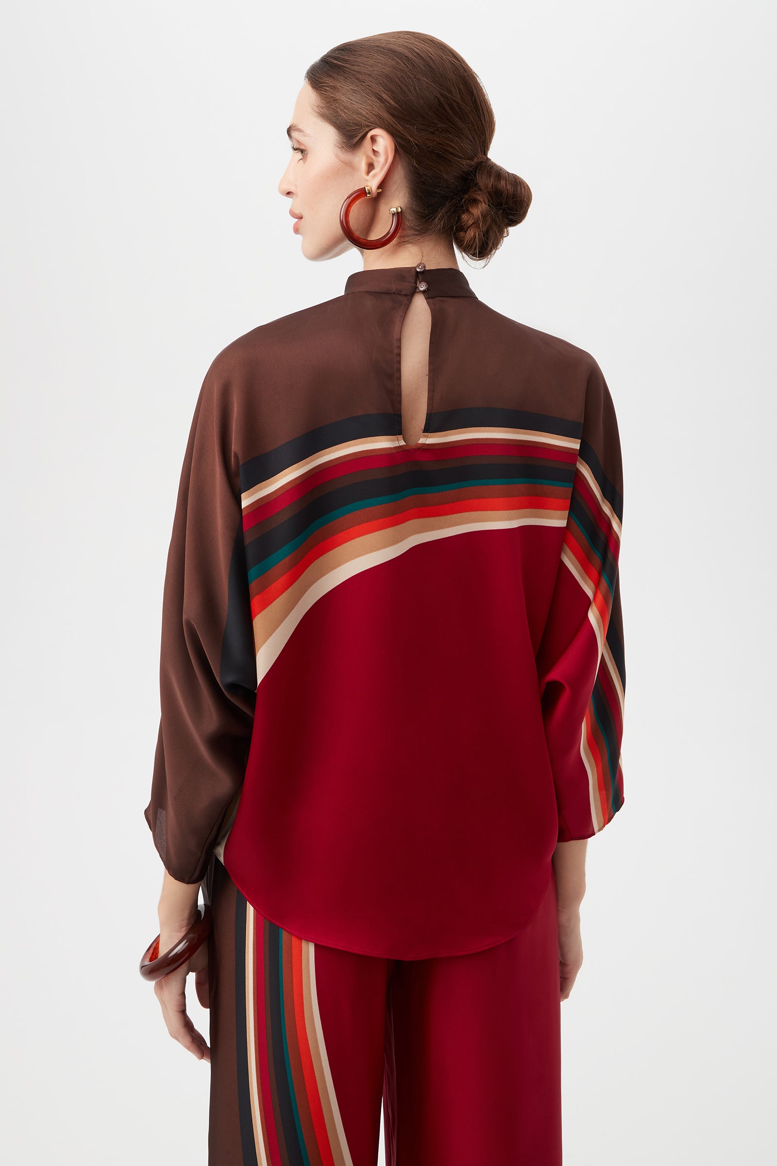 A woman with brown hair in a low bun wears the Kobina Top, a loose, long-sleeved style with red, brown, and multicolored stripes. She faces away, showing the back keyhole detail and large red hoop earrings.