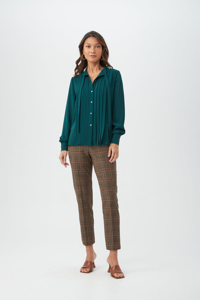 A woman with long, wavy hair and a neutral expression stands against a white background wearing brown plaid Masego Pant, a teal pleated long-sleeve blouse, and brown heeled sandals.