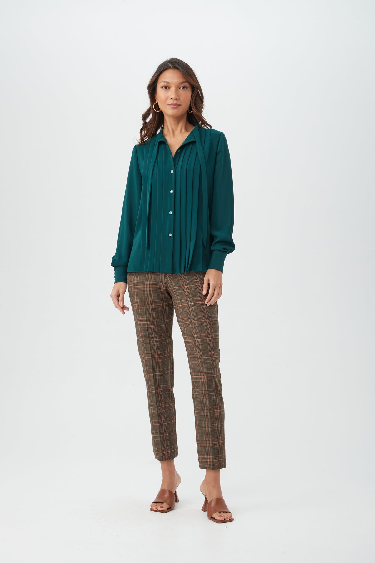 A woman with long, wavy hair and a neutral expression stands against a white background wearing brown plaid Masego Pant, a teal pleated long-sleeve blouse, and brown heeled sandals.