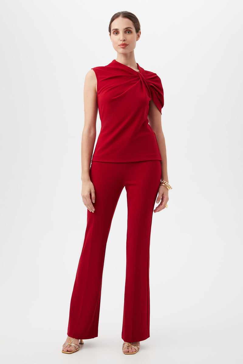 A woman poses against a white background wearing the Jacoba Crepe Knit Tailored Bootcut Pant in red, paired with a sleeveless draped top, gold bracelets, and gold open-toe heels. Her hair is pulled back for a polished look.