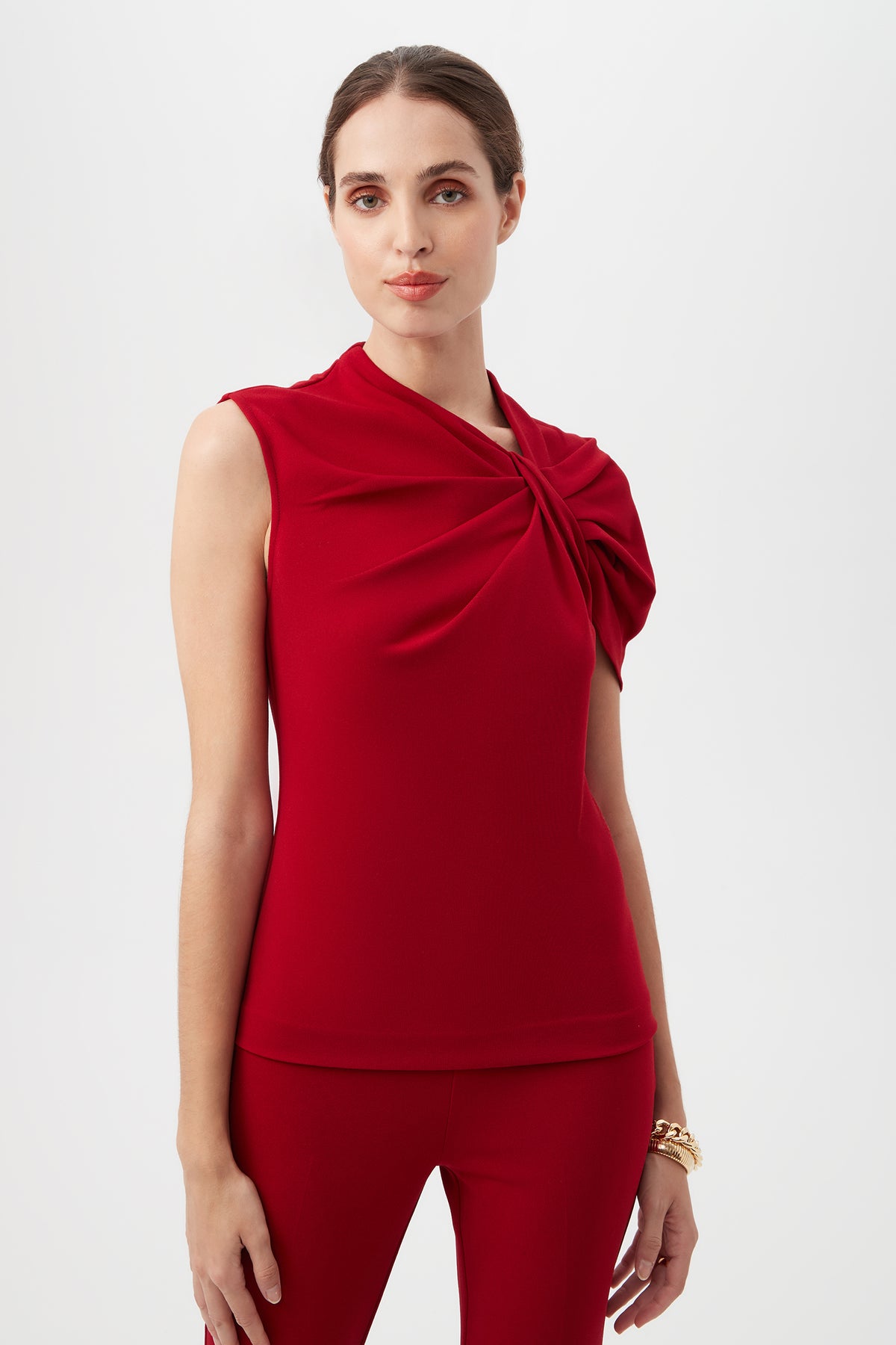 A woman stands against a white background wearing the Keshi Asymmetrical Sleeve Top in ROSELLE RED, featuring a shoulder twist detail, paired with matching red pants.