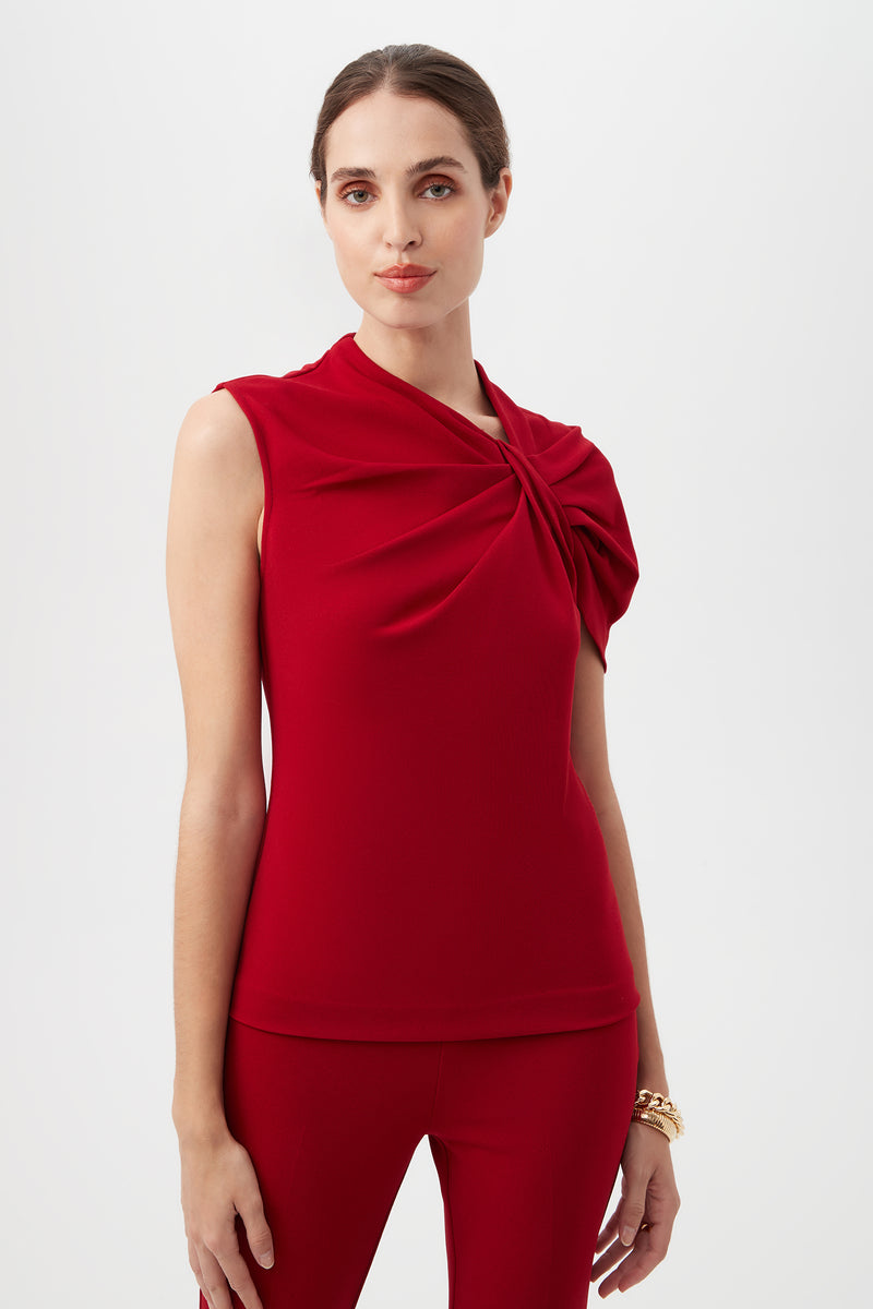 A woman stands against a white background wearing the Keshi Asymmetrical Sleeve Top in ROSELLE RED, featuring a shoulder twist detail, paired with matching red pants.