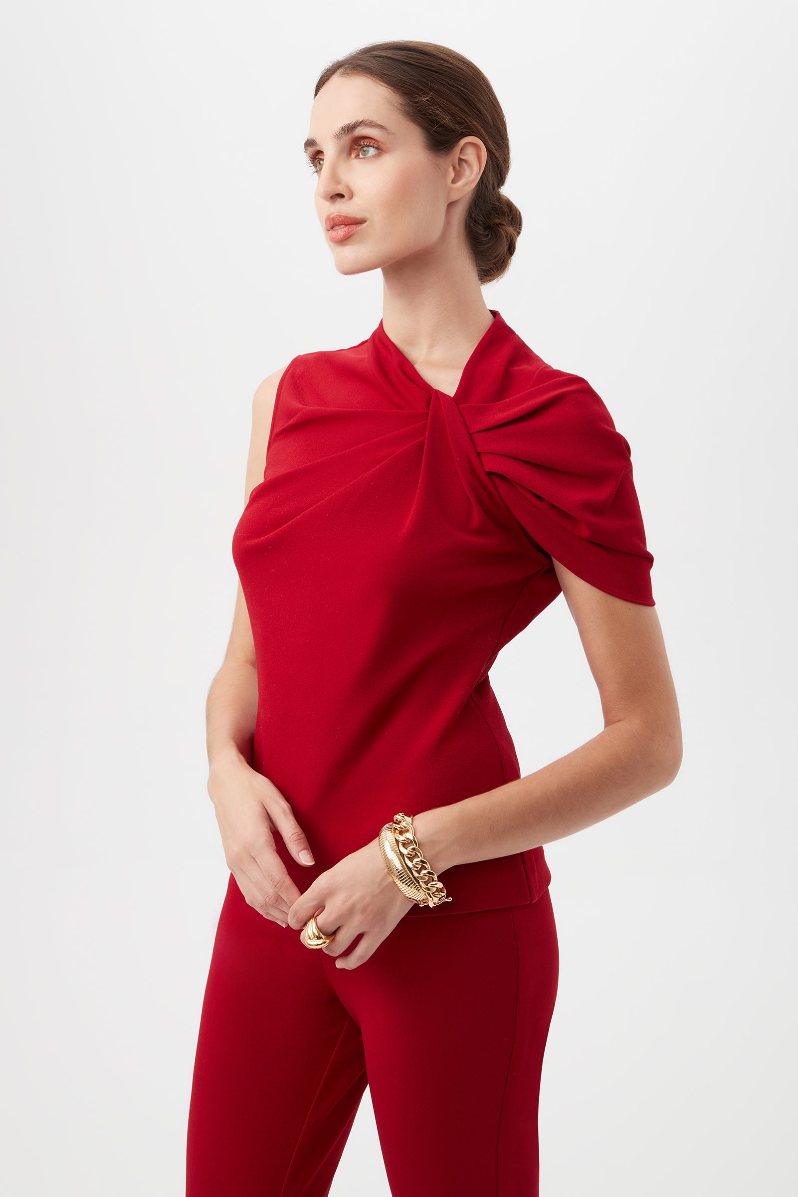 A woman with light skin and brown hair in a low bun wears the Keshi Asymmetrical Sleeve Top With Shoulder Twist Detail in red and matching pants, accessorized with gold bracelets and rings, standing against a plain backdrop.