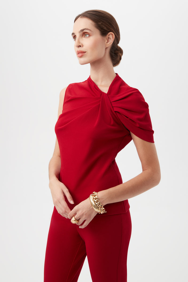 A woman with light skin and brown hair in a low bun wears the Keshi Asymmetrical Sleeve Top With Shoulder Twist Detail in red and matching pants, accessorized with gold bracelets and rings, standing against a plain backdrop.