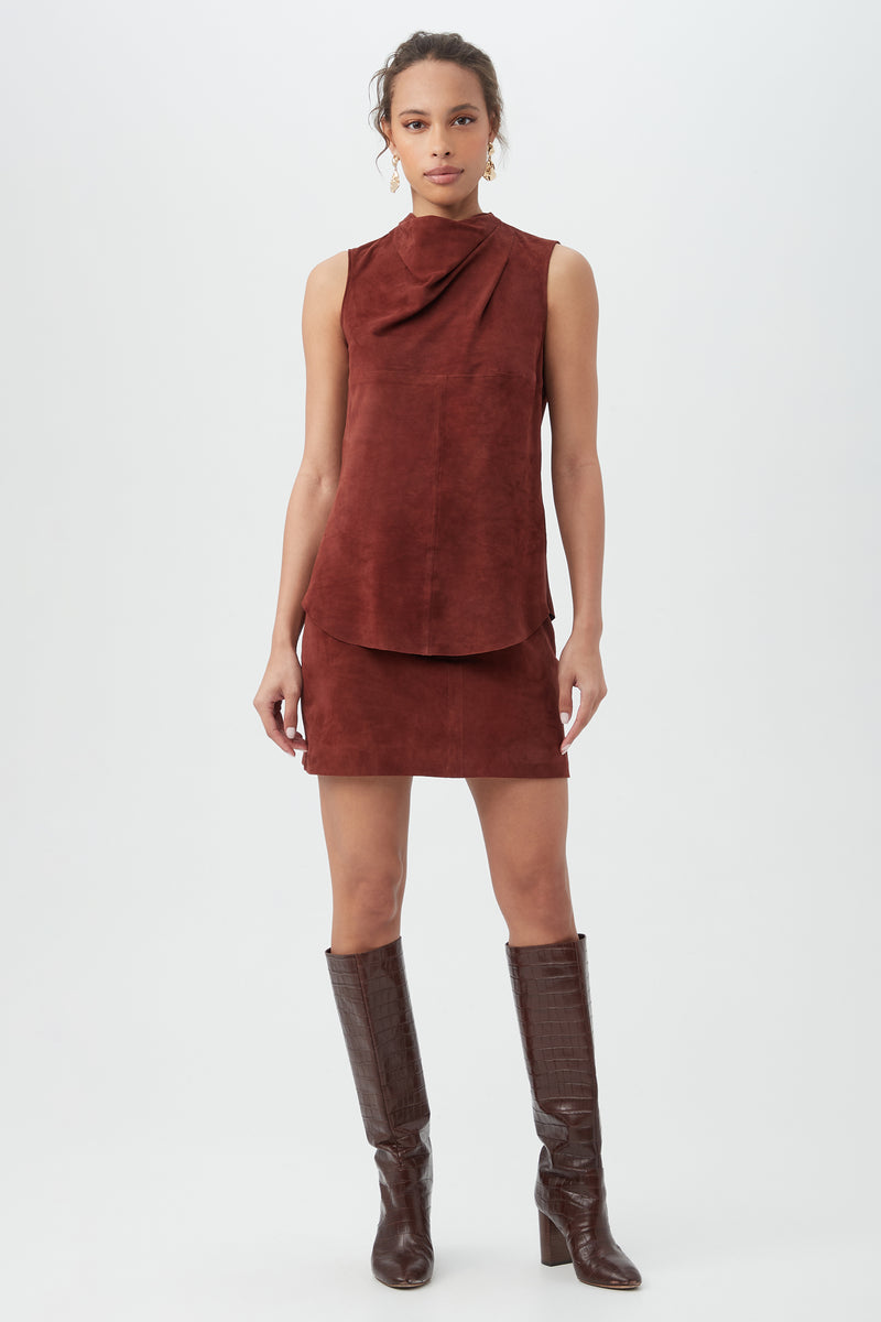 A woman stands against a plain background wearing the Lenaya 2 Top, a sleeveless rust suede piece with a draped neckline, paired with a matching mini skirt and knee-high brown leather boots.