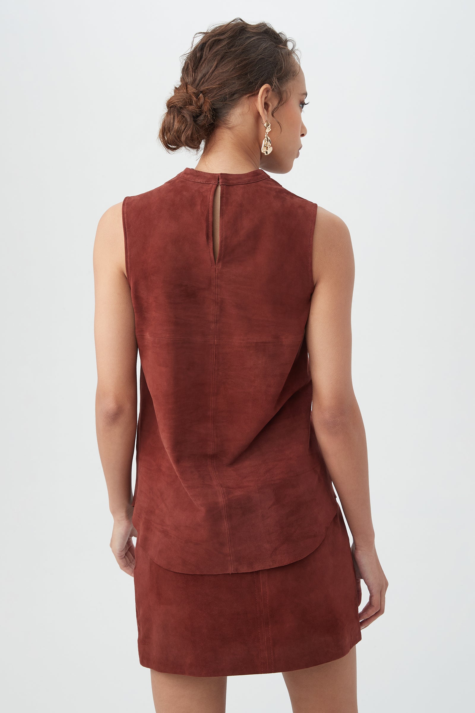 Woman with short curly hair in a low bun, wearing gold earrings and the rust-colored Lenaya 2 Top, shown from the back against a plain light background.