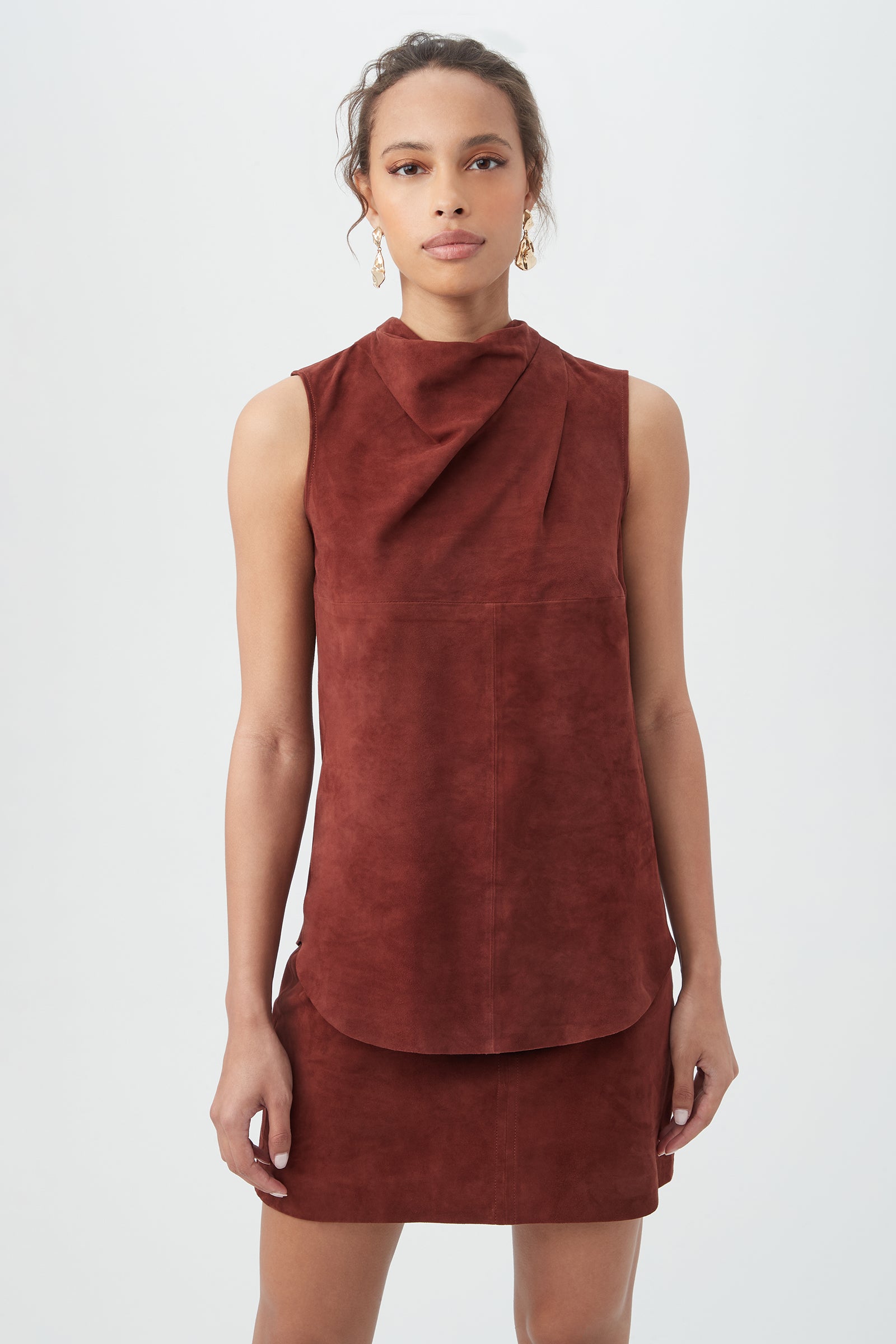 A woman wearing the Lenaya 2 Top in mahogany, featuring a sleeveless design and high draped neckline, stands against a plain white background with earrings on and her hair tied back.