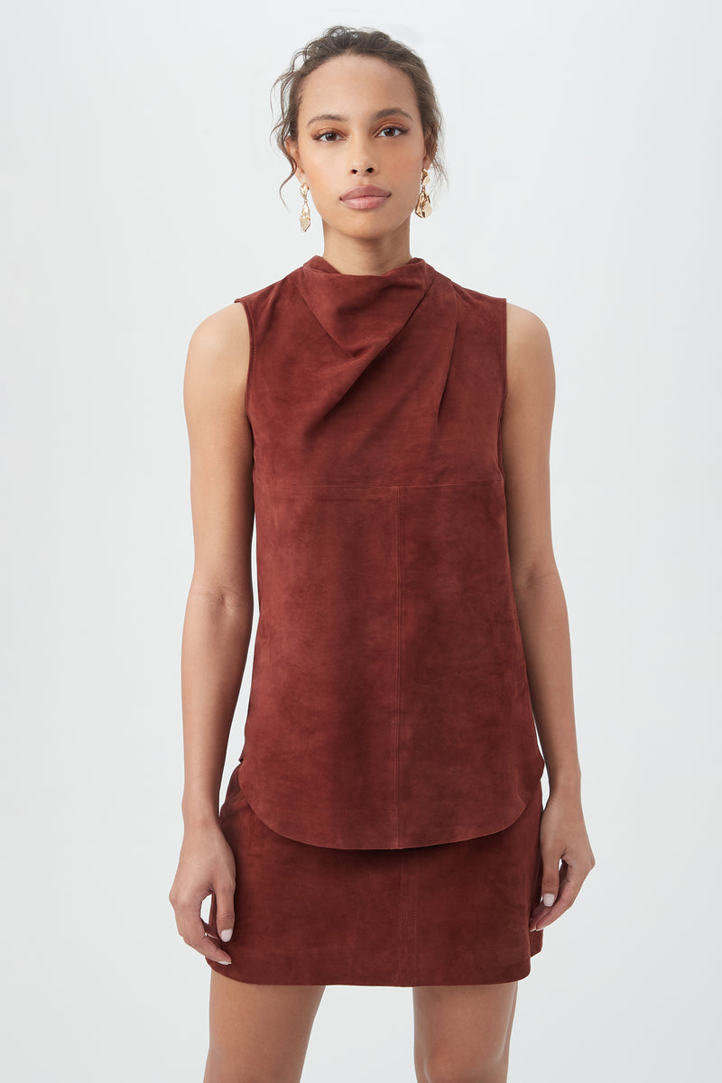 A woman wearing the Lenaya 2 Top in mahogany, featuring a sleeveless design and high draped neckline, stands against a plain white background with earrings on and her hair tied back.