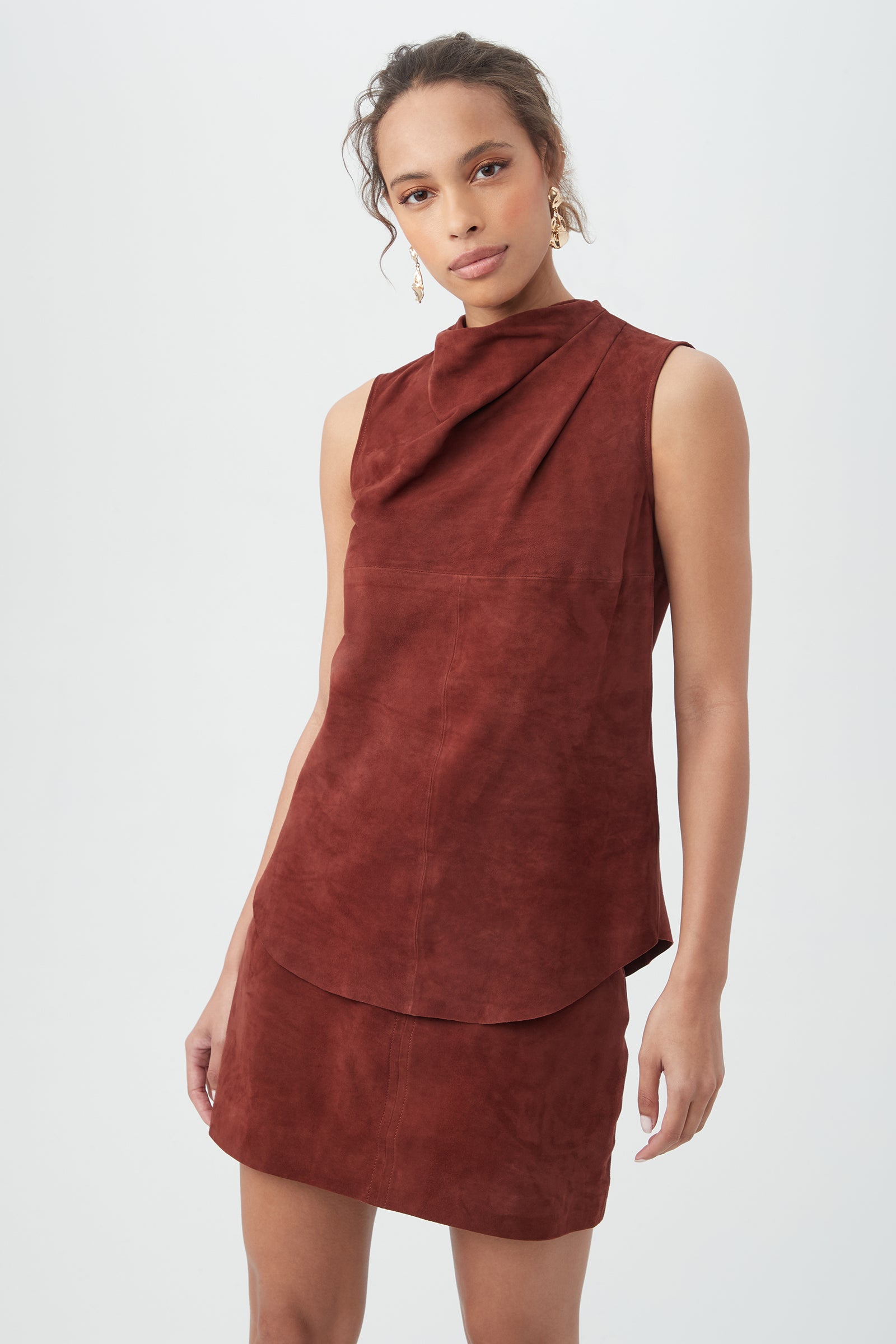 A woman wearing the Lenaya 2 Top, a sleeveless rust suede piece with a draped neckline, and a matching mini skirt stands against a plain white background.