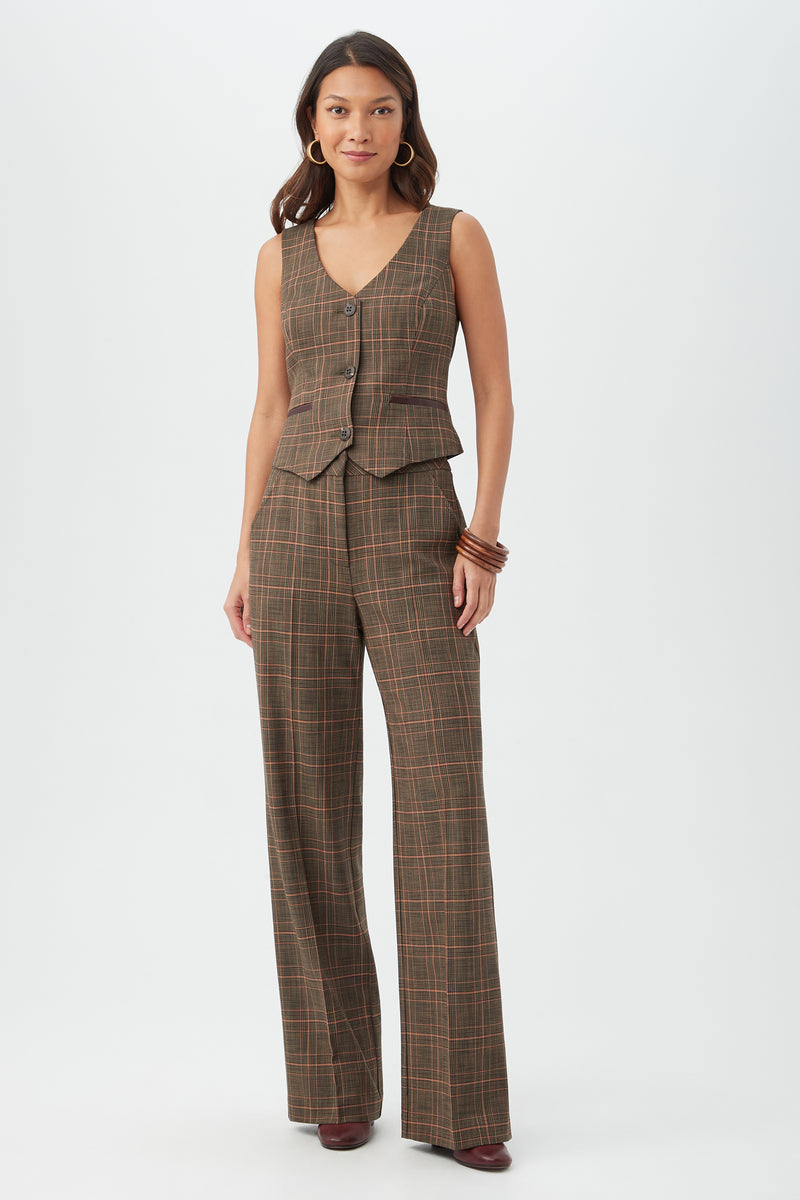 A woman with long hair, hoop earrings, and bangles stands against a white background wearing the Kofi Vest with matching brown plaid wide-leg pants and maroon shoes.