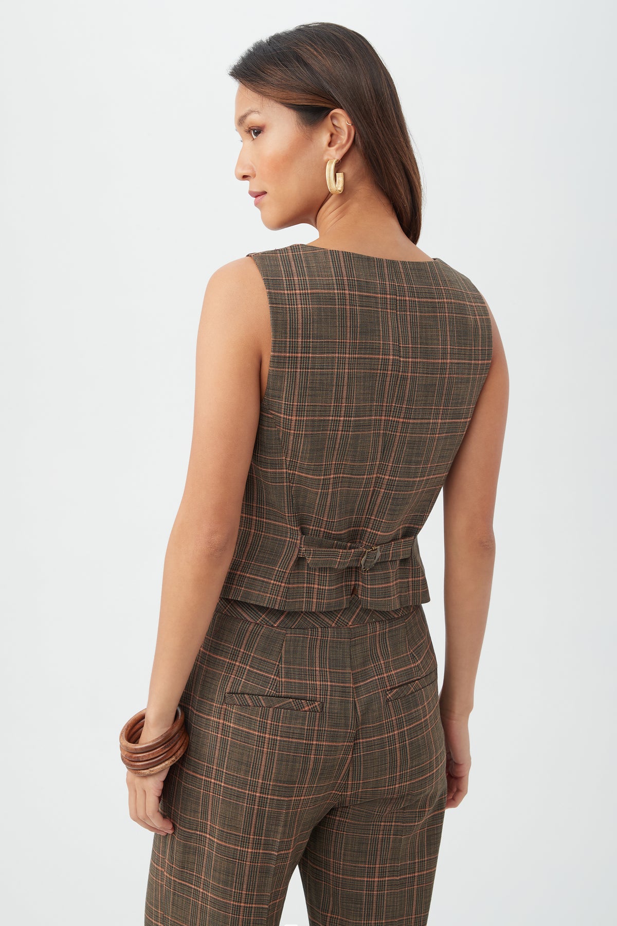 A woman with long, straight hair wears the sleeveless Kofi Vest in brown plaid with matching pants, accessorized with gold hoop earrings and chunky bracelets, standing against a plain light background.