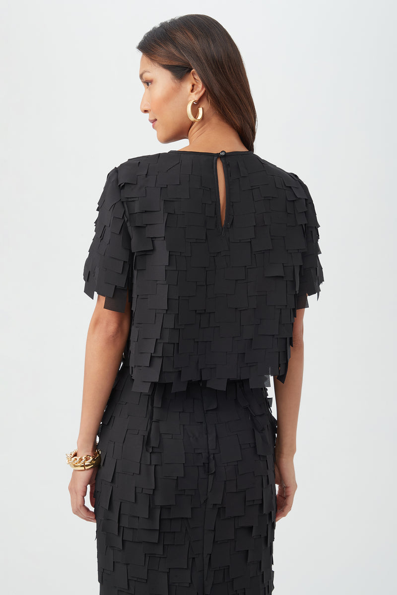 A woman with long brown hair, seen from behind, wears the Serilda Short-Sleeve Top in black flutter chiffon with layered squares, short sleeves, a top button closure, and gold jewelry.