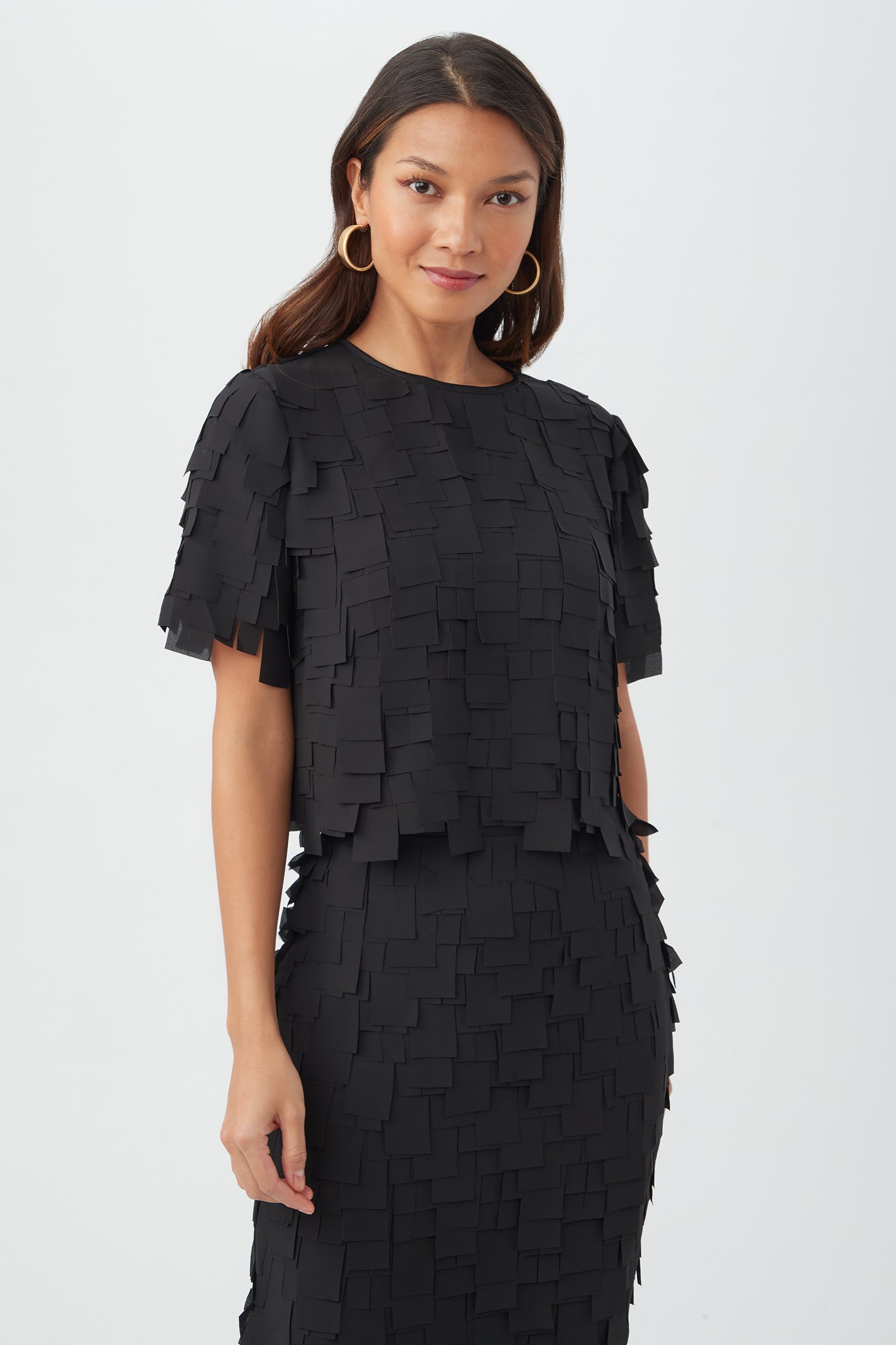 A woman in the black Serilda Short-Sleeve Top - Flutter Chiffon, styled with large gold hoop earrings and long wavy hair, stands against a plain white background, smiling slightly.