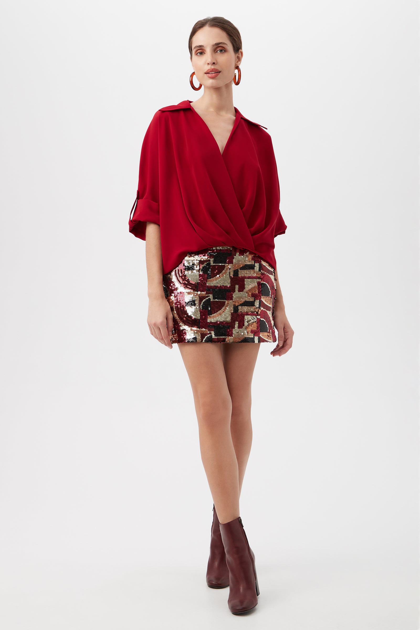 A woman stands against a white background wearing a loose deep red blouse, maroon ankle boots, large red hoop earrings, and the Rico Galerie Arte Sequin Mini Skirt.