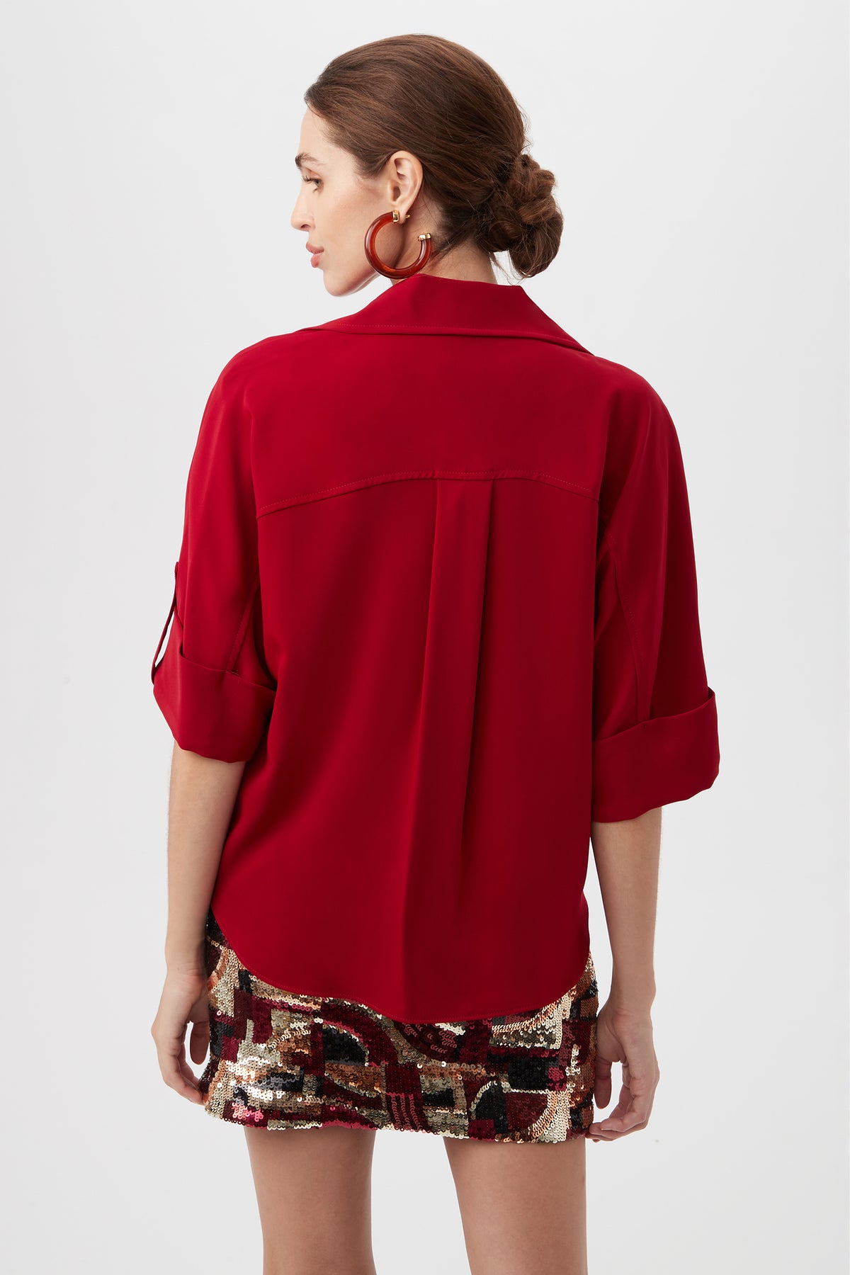 A woman faces away, wearing the Otto Top—a bright red long-sleeve blouse with rolled-up sleeves—paired with a shiny gold, black, and red patterned mini skirt; her hair is in a low bun and she has large hoop earrings.
