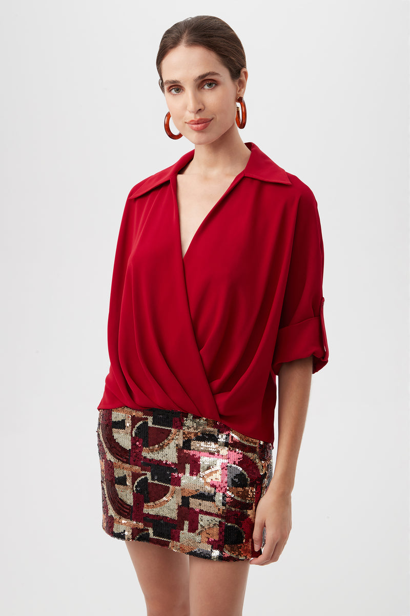 A woman with brown hair pulled back and large hoop earrings wears the Otto Top—a red wrap blouse with rolled sleeves—and a short, patterned sequin skirt, standing against a plain background with a neutral expression.