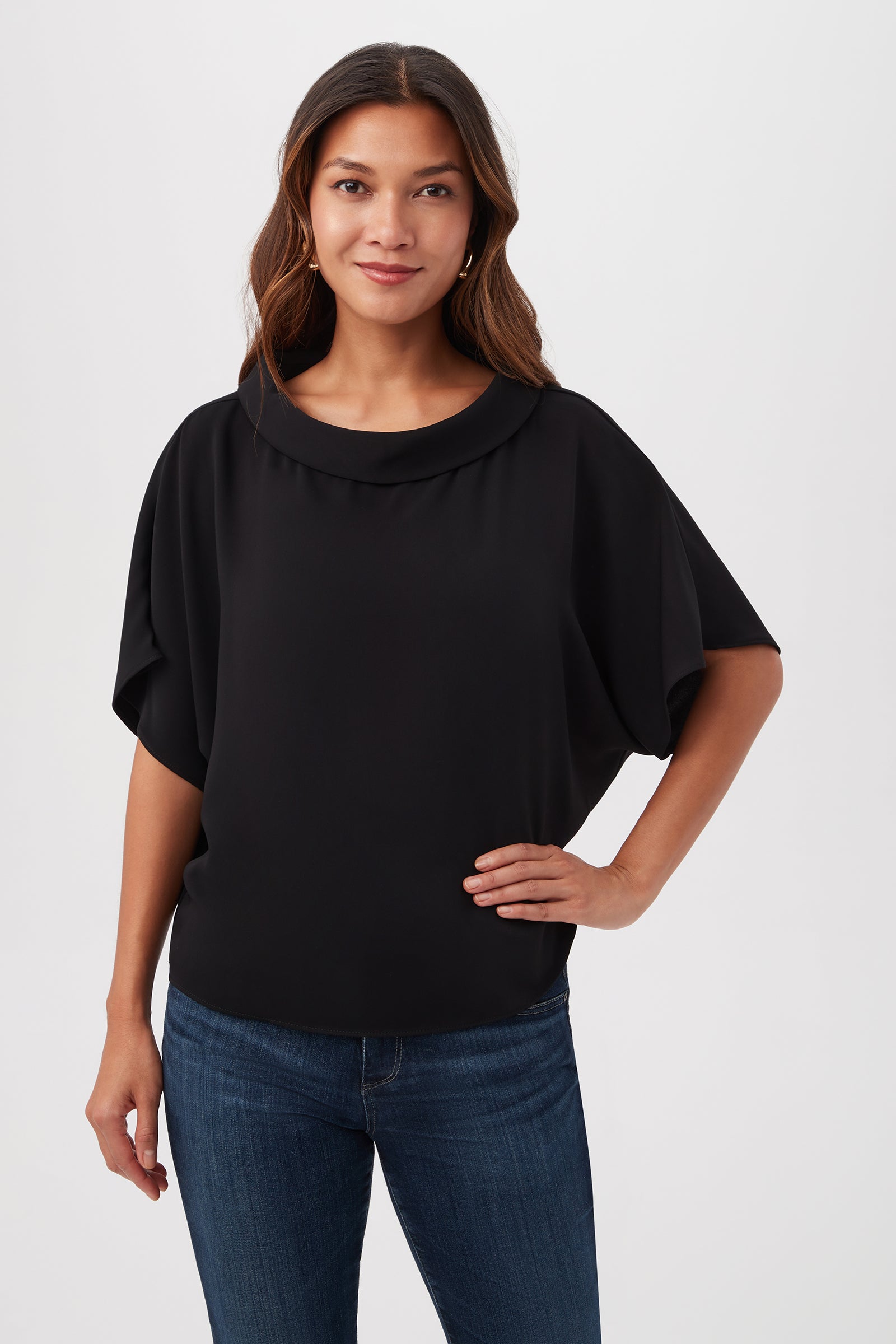 A woman with long brown hair wears the Honeysuckle Boatneck Dolman-Sleeve Top in BLACK and dark blue jeans. She stands against a plain white background, smiling slightly with one hand on her hip.