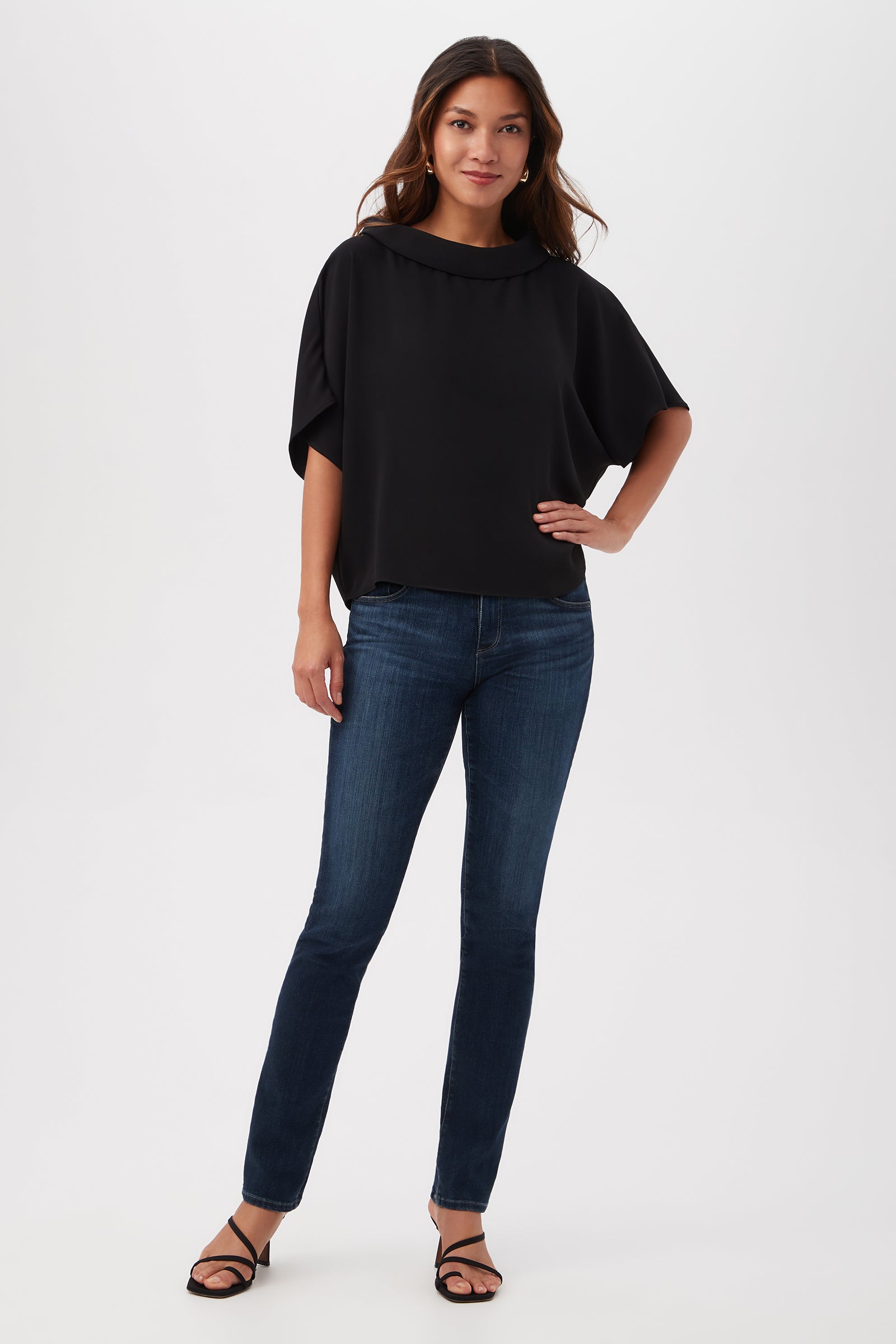 A woman with long brown hair stands against a white background, wearing the Honeysuckle Boatneck Dolman-Sleeve Top in black, paired with dark blue skinny jeans and black strappy heels. She poses confidently with one hand on her hip.