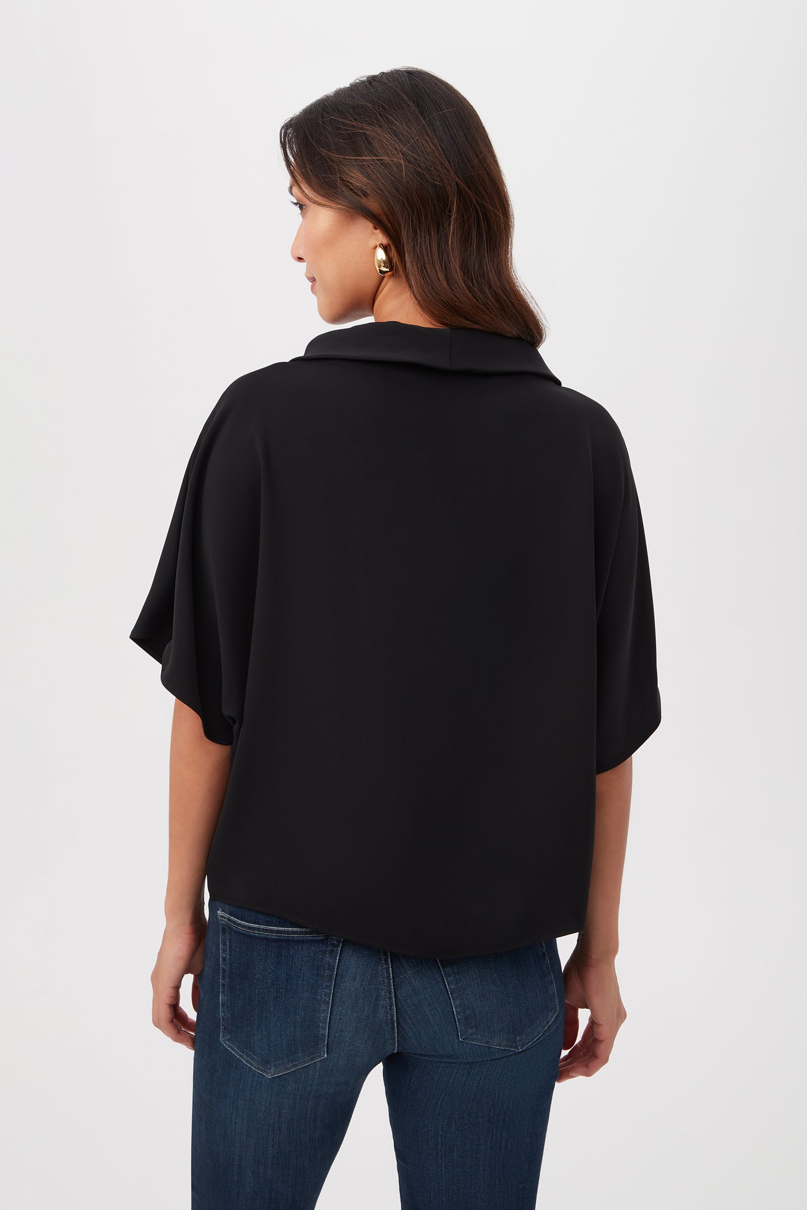 Woman with wavy brown hair wearing the Honeysuckle Boatneck Dolman-Sleeve Top in black and blue jeans, standing with her back to the camera against a plain light background. She pairs the look with large gold earrings.