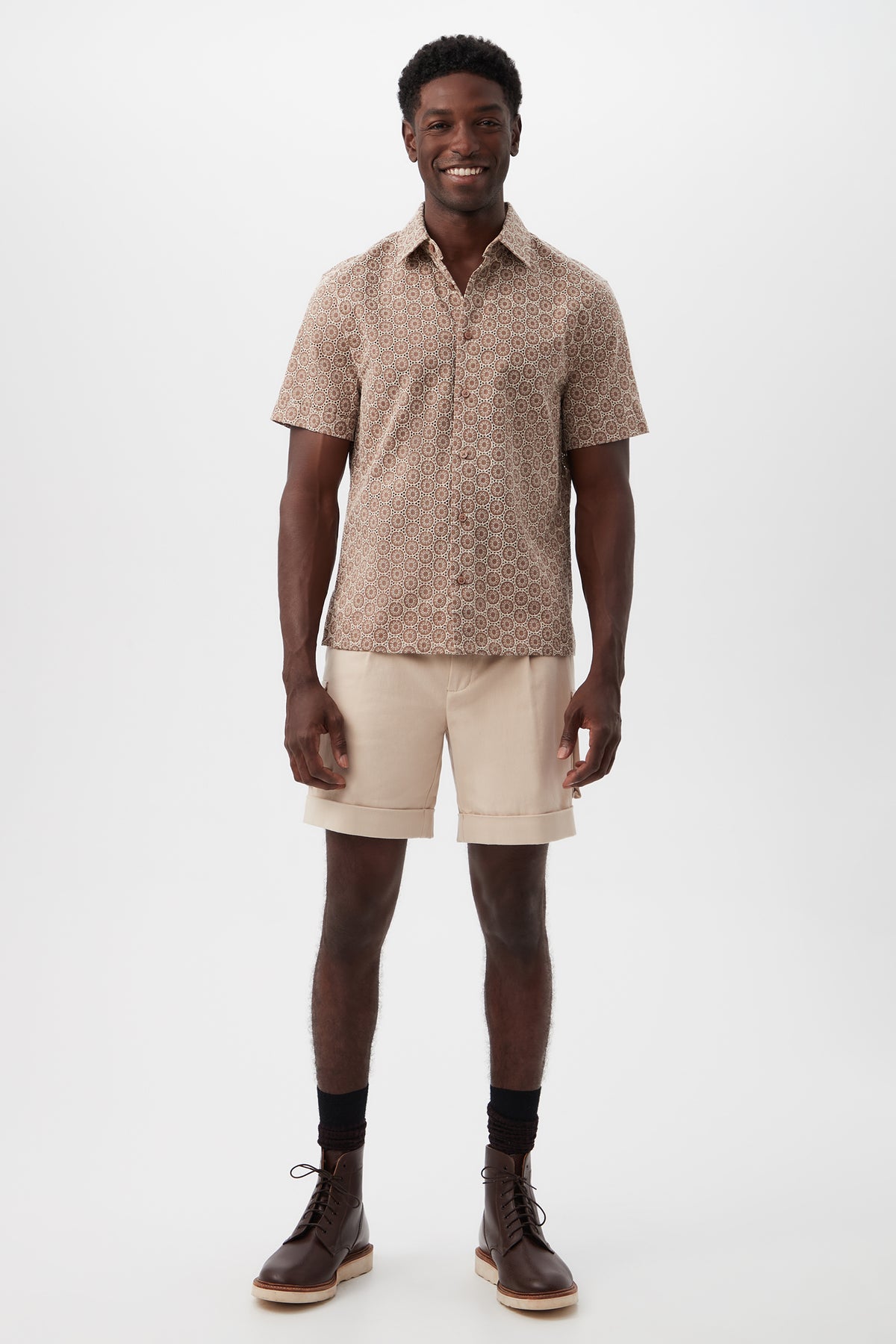A man stands smiling in a short-sleeve patterned beige shirt, black socks, brown boots, and Westlake Cargo Short - Brushed Cotton Twill in light beige, set against a plain white background.