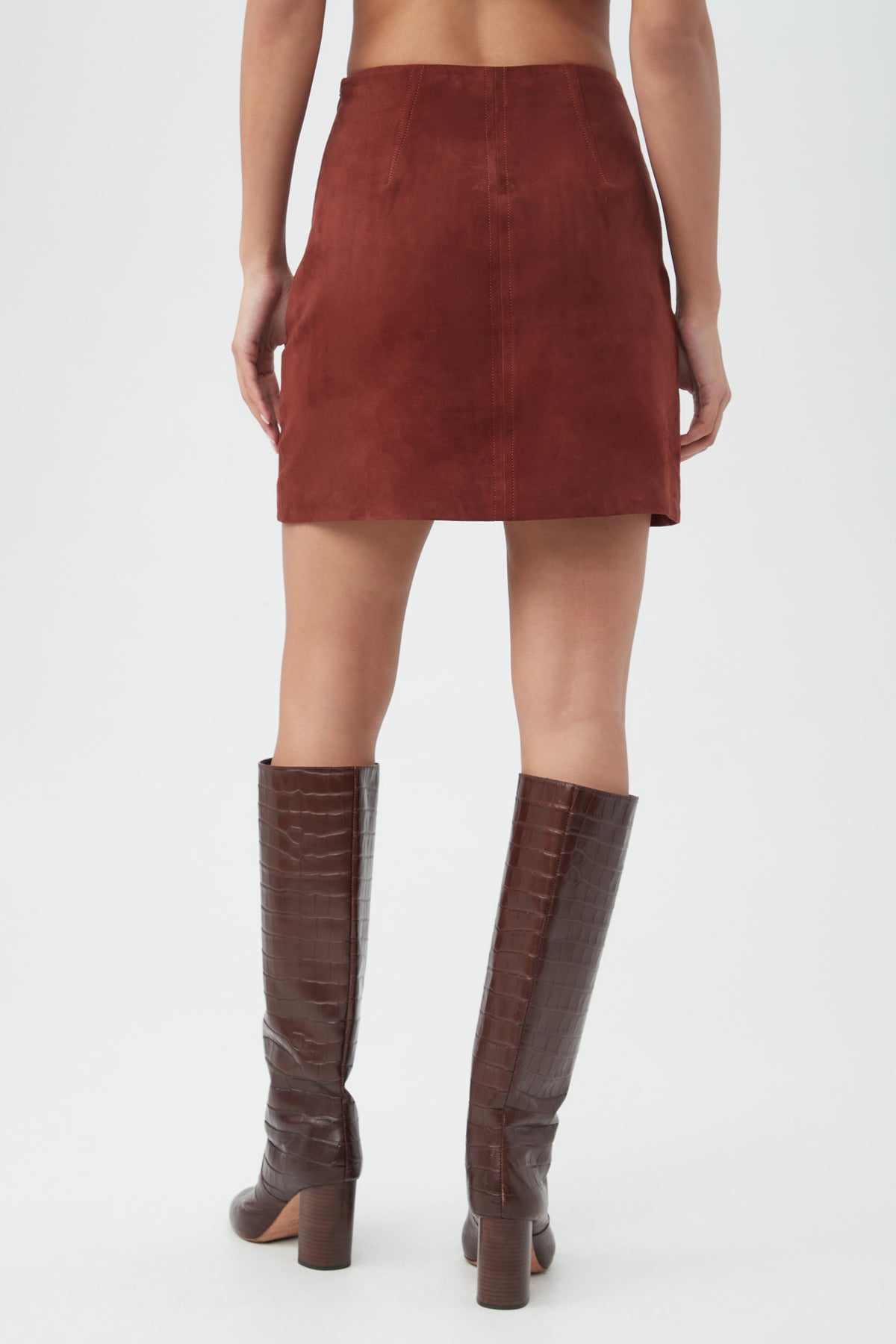 A person is shown from the back wearing the High Waisted Rico 2 Skirt in reddish-brown suede and knee-high, brown, crocodile-textured boots against a plain white background.