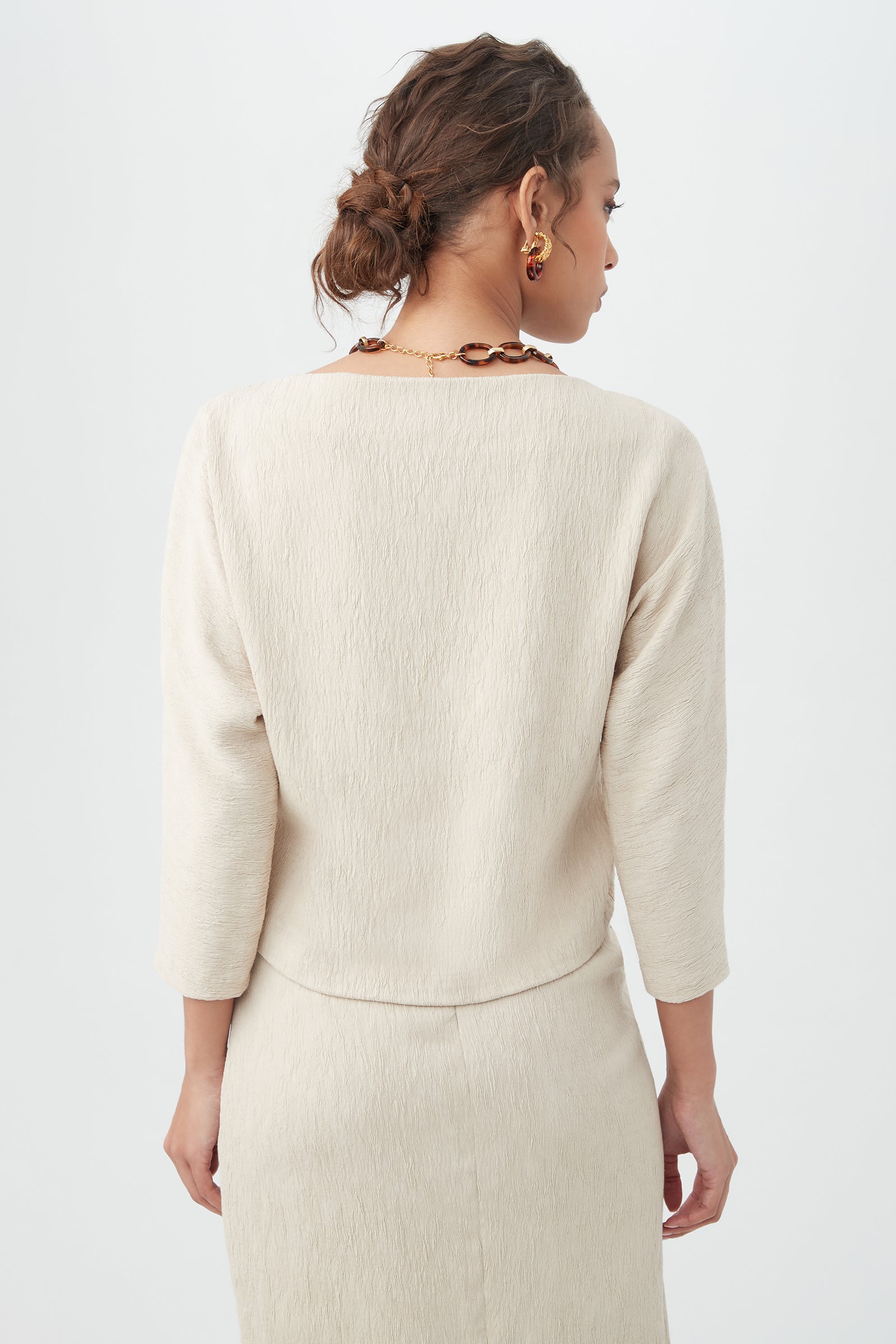A woman with wavy brown hair in an updo is shown from the back wearing the cream-colored, textured Imma Top and matching skirt, paired with gold earrings and a chunky chain necklace against a plain white background.