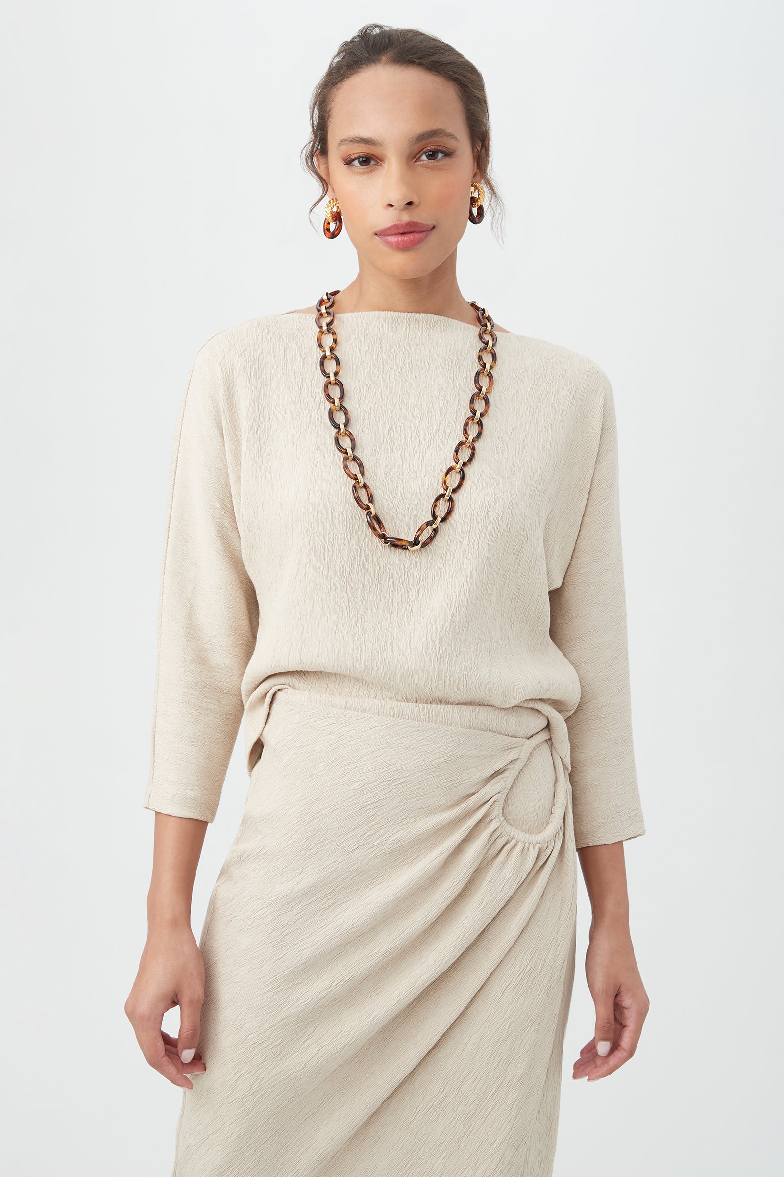 A woman wears the Imma Top in Sahara Stone with a matching skirt featuring a side knot, accessorized with a chunky tortoiseshell necklace and hoop earrings, standing against a plain light background.