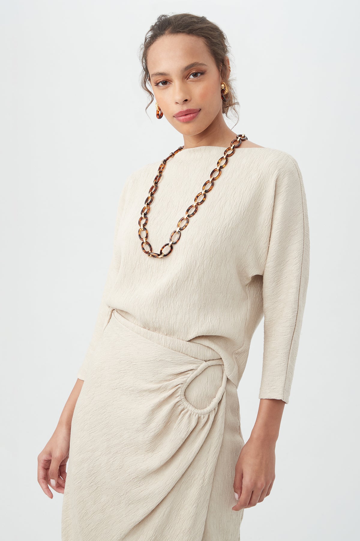 A woman wears the Imma Top—a beige, textured long-sleeve top paired with a matching skirt featuring gathered detail and a circular accent—styled with a chunky brown tortoiseshell necklace and gold hoop earrings.