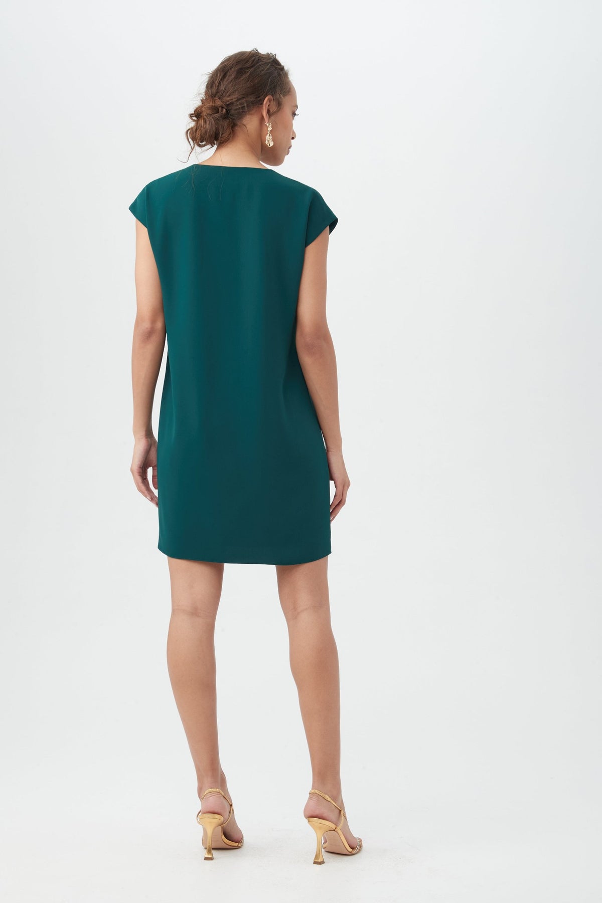 A woman faces away, wearing the Zula Dress—a short-sleeved, knee-length teal dress—with beige high-heeled sandals against a plain white background.