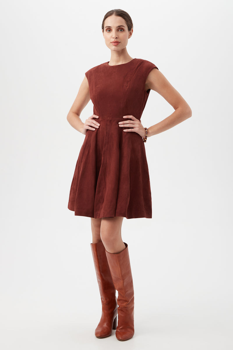 A woman poses with hands on hips in the Sabi Dress, a sleeveless maroon dress with a flared skirt, styled with brown knee-high boots. Her hair is pulled back and she stands against a white background.