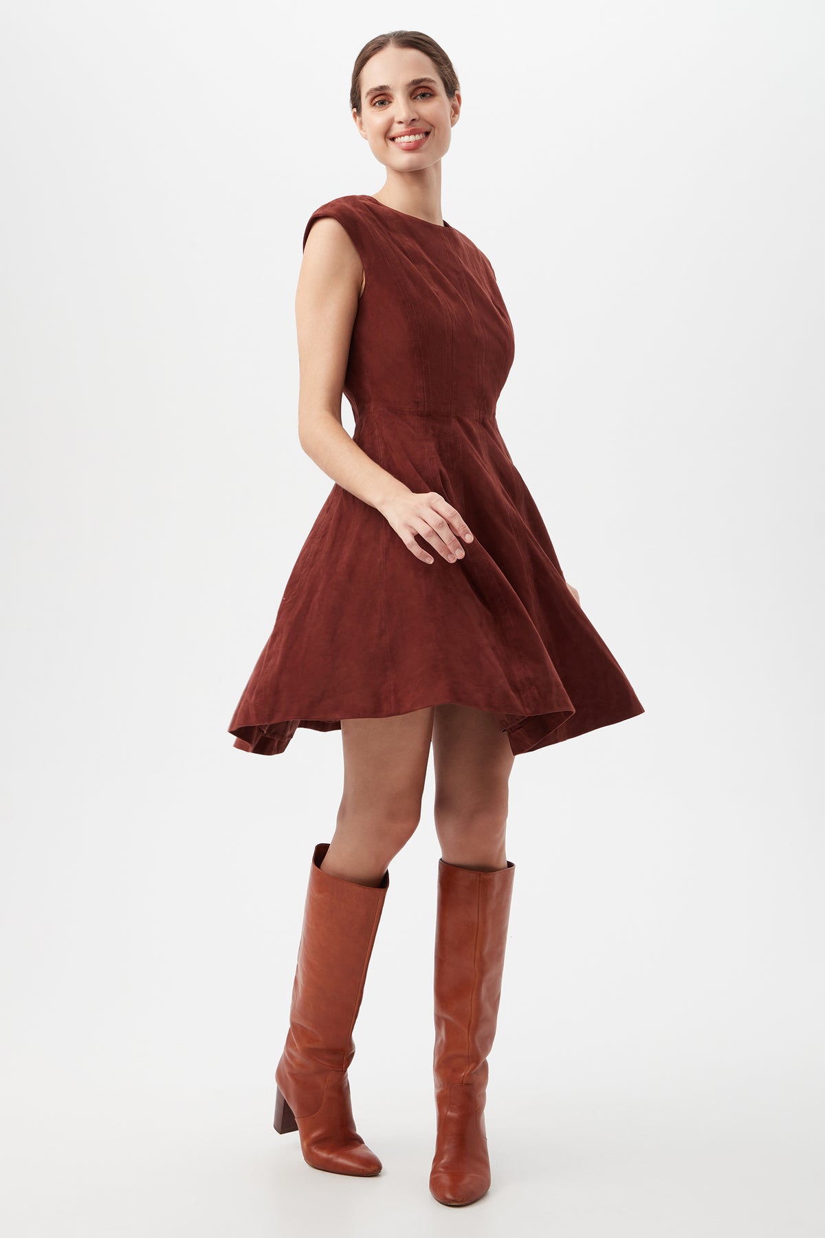 A woman smiles and poses in the Sabi Dress, a sleeveless dark red A-line with a flared skirt, paired with tall brown boots, against a plain white background.