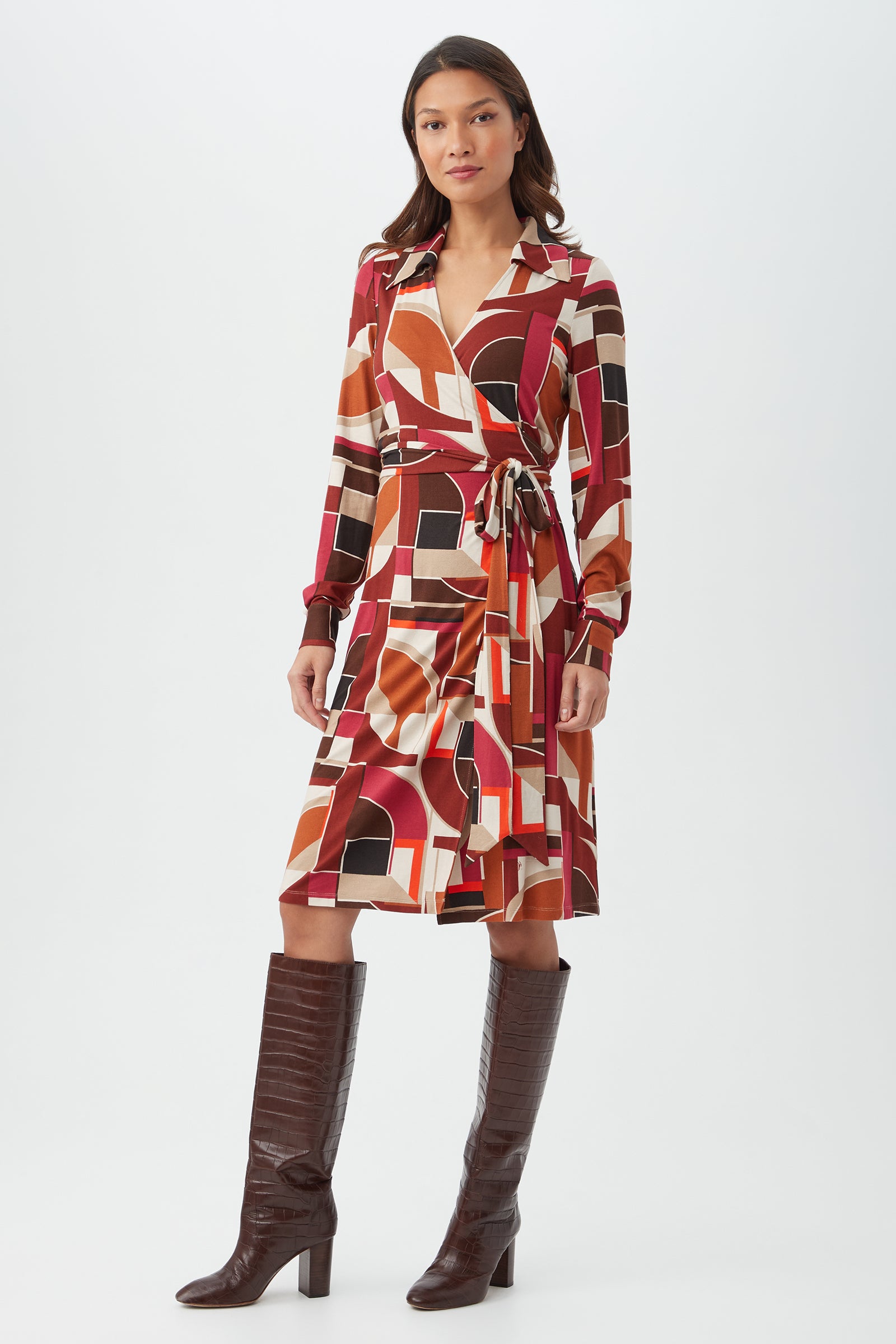 A woman stands against a white background wearing the knee-length, long-sleeve Makeba Dress in a multi geometric print, paired with tall brown heeled boots.