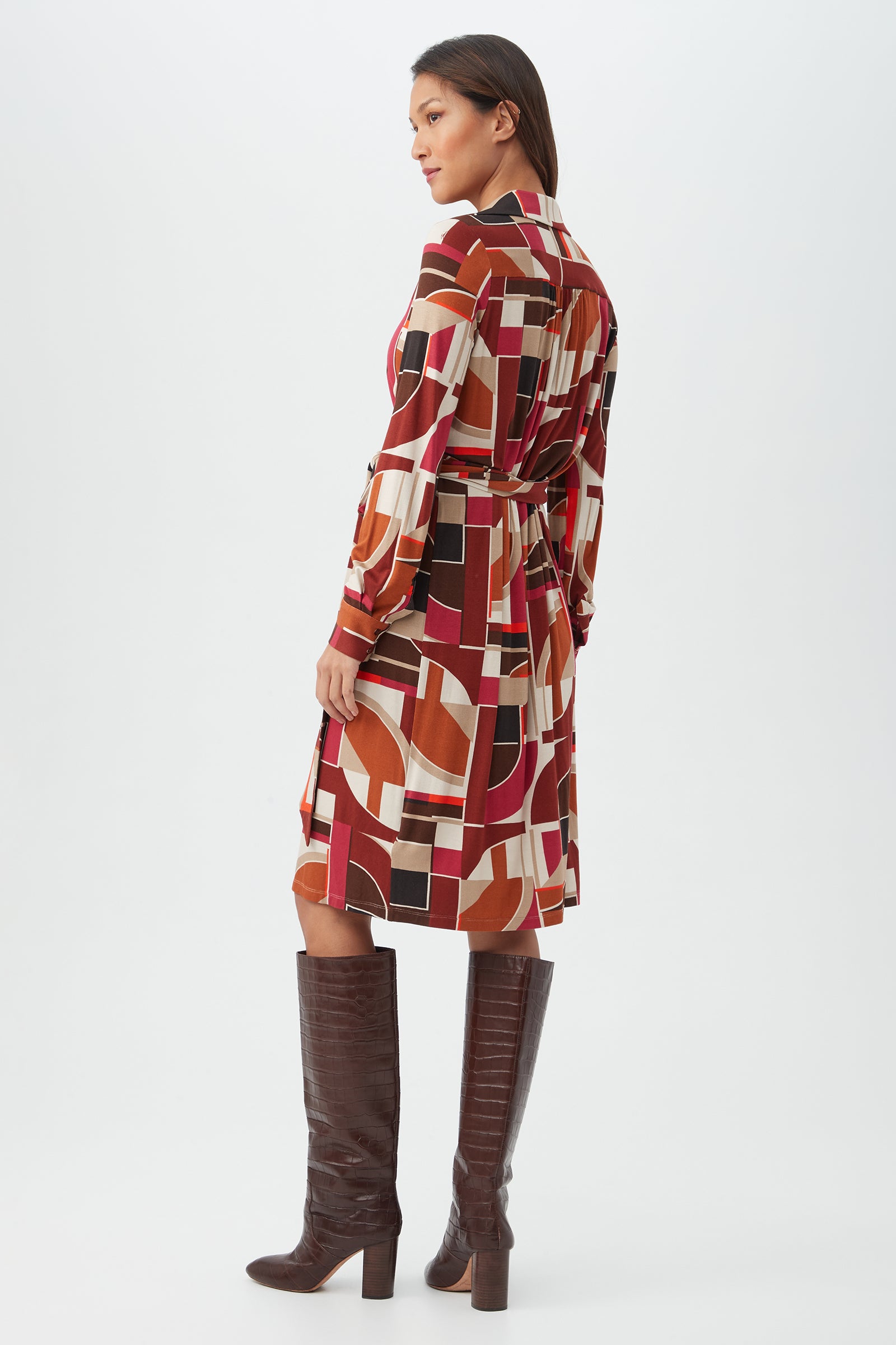 A woman stands sideways in the Makeba Dress—a knee-length, long-sleeve dress with a bold red, orange, cream, and black geometric pattern—paired with tall dark brown heeled boots against a plain white background.