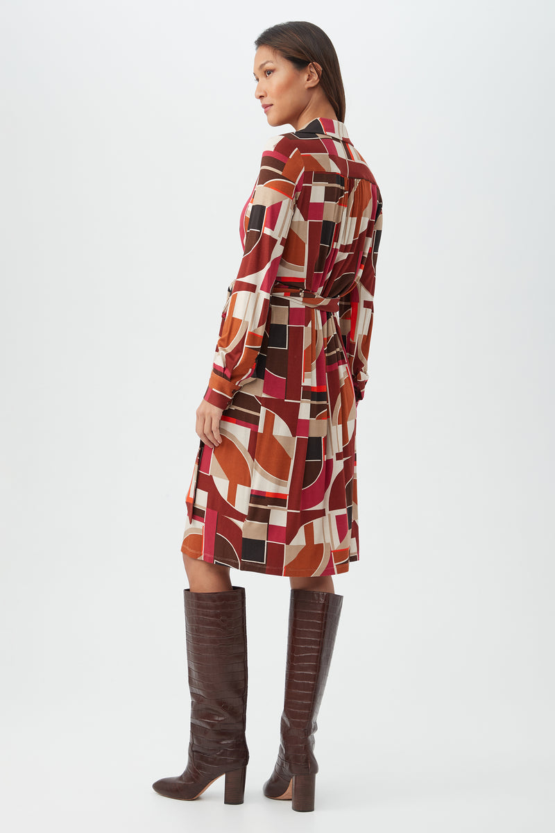 A woman stands sideways in the Makeba Dress—a knee-length, long-sleeve dress with a bold red, orange, cream, and black geometric pattern—paired with tall dark brown heeled boots against a plain white background.