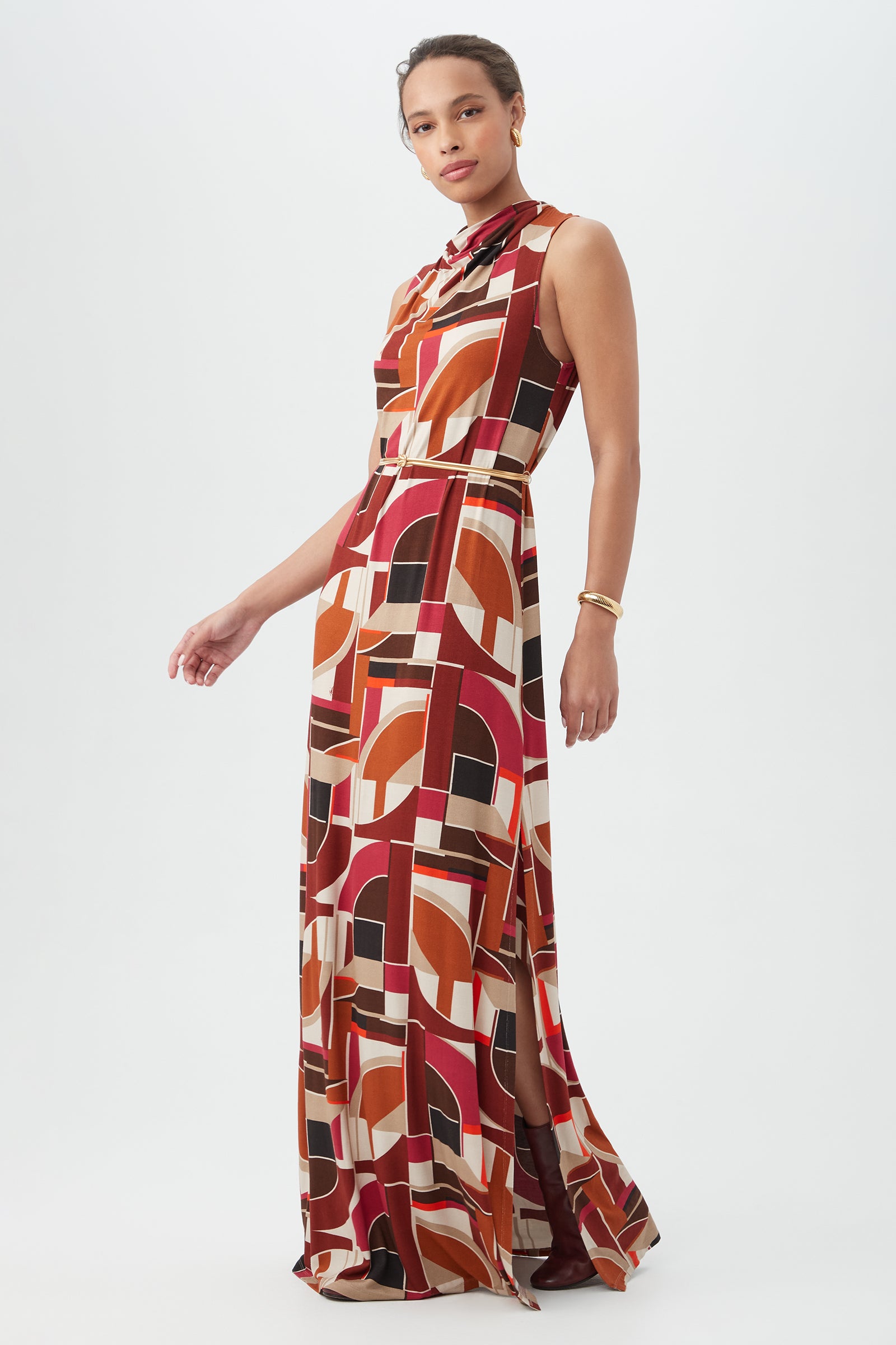A woman with her hair pulled back and a bracelet stands against a white background, wearing the Kia Dress—a sleeveless, floor-length dress featuring a bold geometric pattern in red, orange, black, and beige.