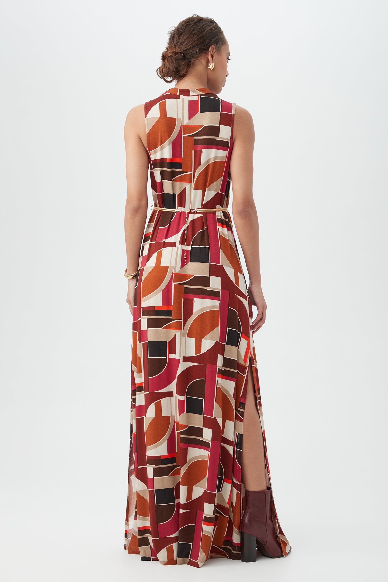 A woman stands facing away, wearing the Kia Dress—a sleeveless, floor-length style with a bold geometric pattern in red, orange, beige, and black, featuring a side slit. She pairs it with brown ankle boots.