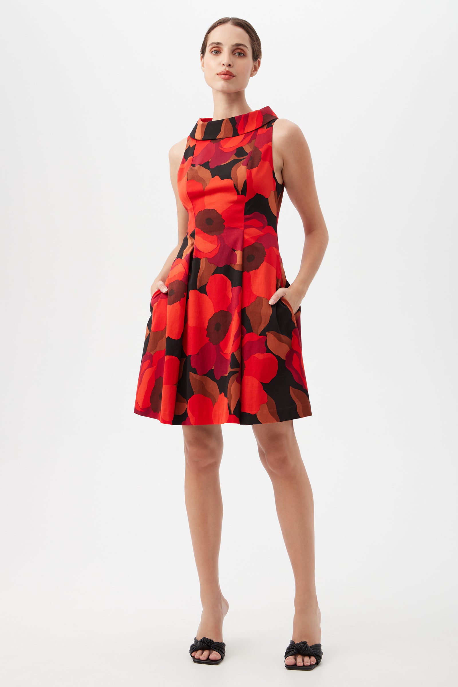 A woman wears the Sabar Red Floral Sleeveless Cocktail Dress in Multi, featuring a knee-length cut and bold floral pattern. She stands against a white background, hands in the dress pockets, paired with black sandals.