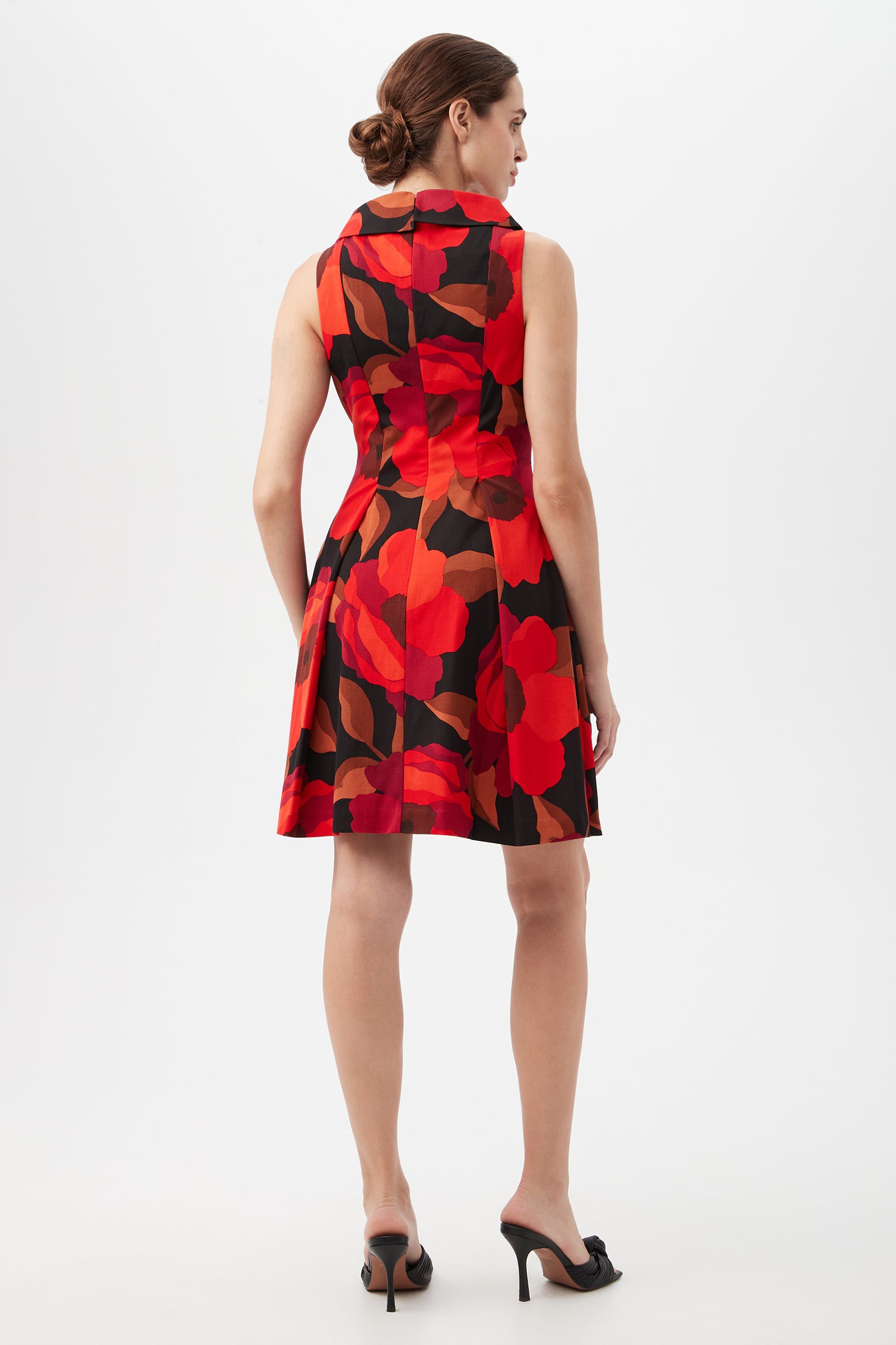 A woman with her hair in a low bun wears the Sabar Red Floral Sleeveless Cocktail Dress, featuring a bold red and black floral pattern and knee-length cut. She pairs it with black heeled shoes against a plain white background.