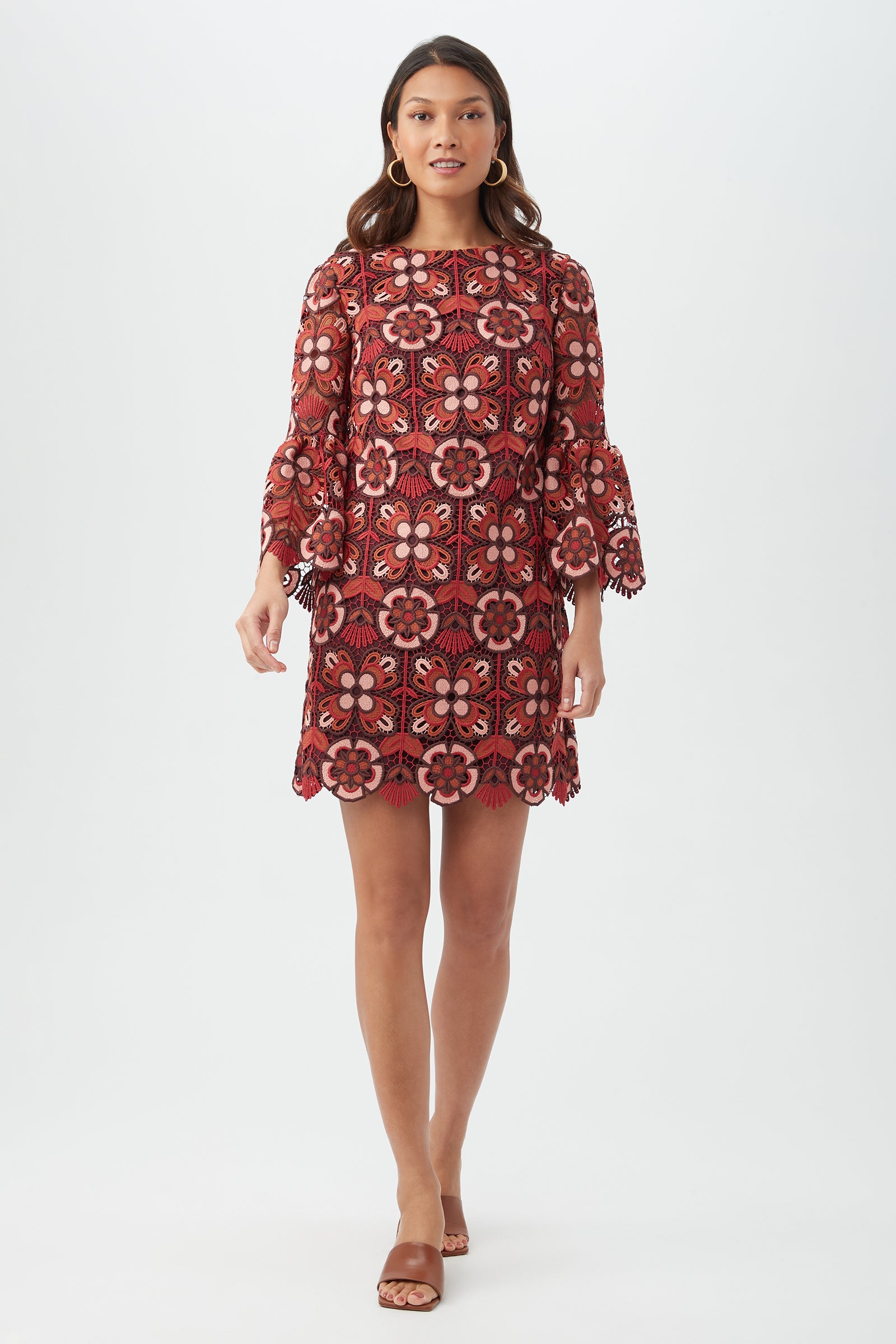 A woman with long wavy hair stands against a white background wearing the Smolder Dress in Multi, a knee-length dress with long sleeves, floral pattern, and scalloped edges. She completes the look with brown sandals.