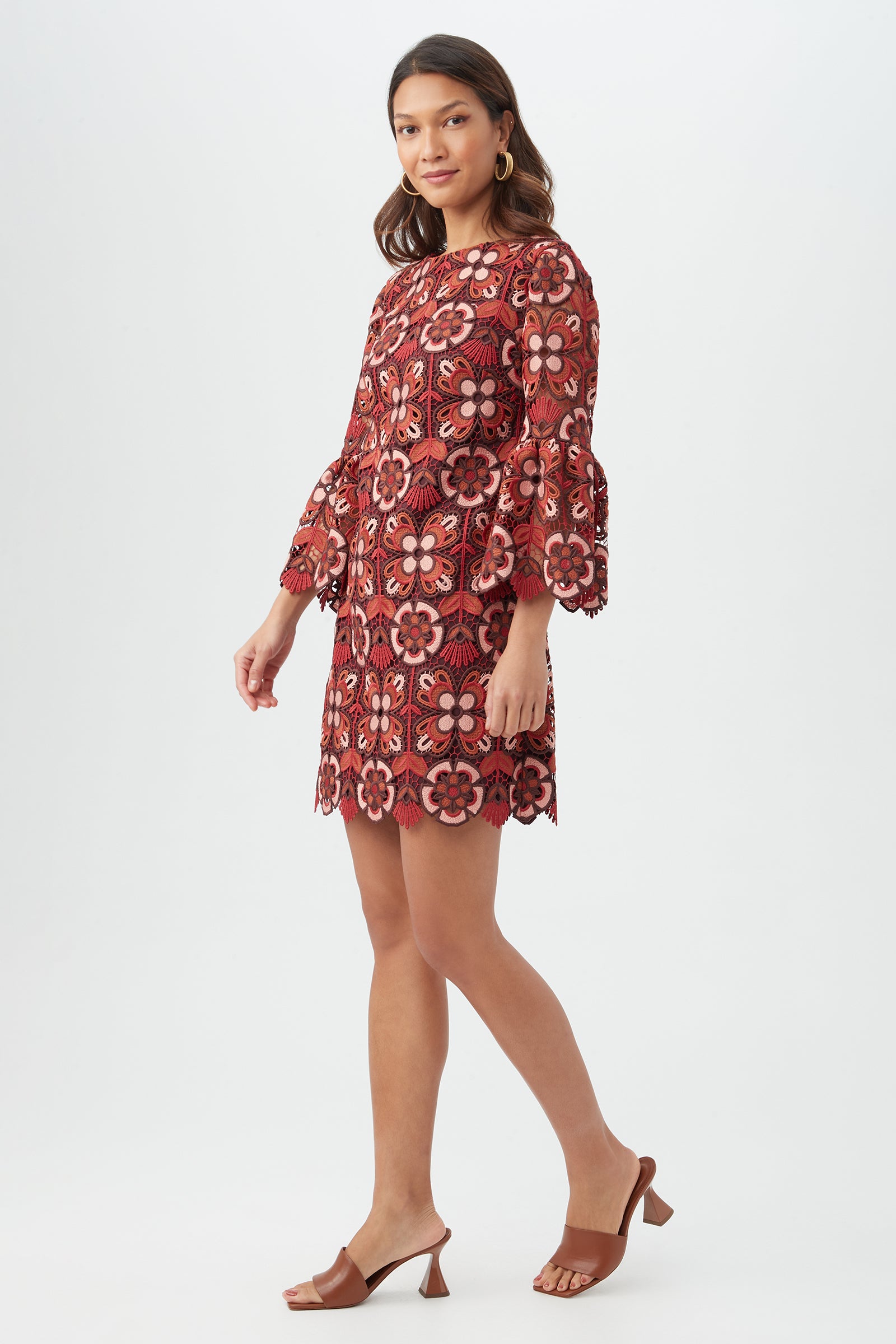 A woman models the Smolder Dress, a red floral lace dress with bell sleeves, paired with brown heeled sandals, standing against a white background and smiling softly over her shoulder.