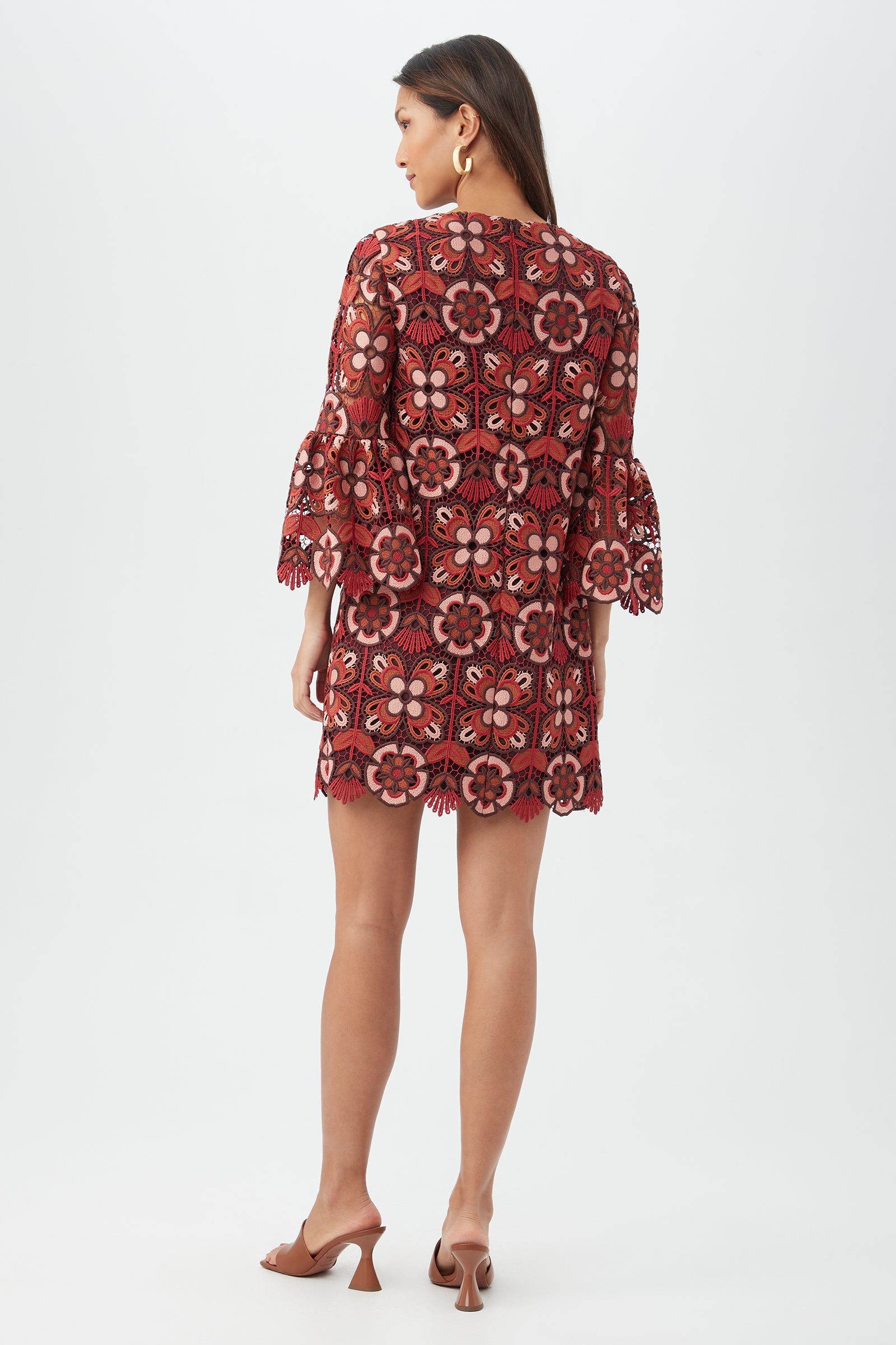 A woman with long brown hair and hoop earrings faces away, modeling the Smolder Dress—a short, loose-fitting dress with large red and pink floral lace patterns. She pairs it with brown heeled sandals against a plain white background.