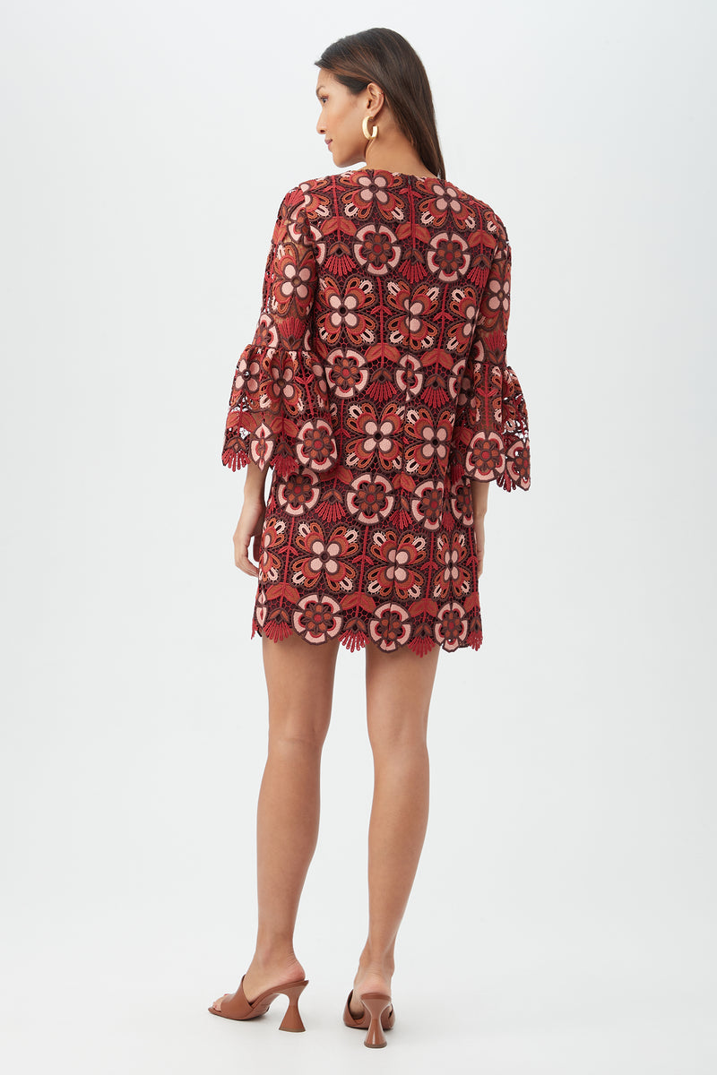A woman with long brown hair and hoop earrings faces away, modeling the Smolder Dress—a short, loose-fitting dress with large red and pink floral lace patterns. She pairs it with brown heeled sandals against a plain white background.