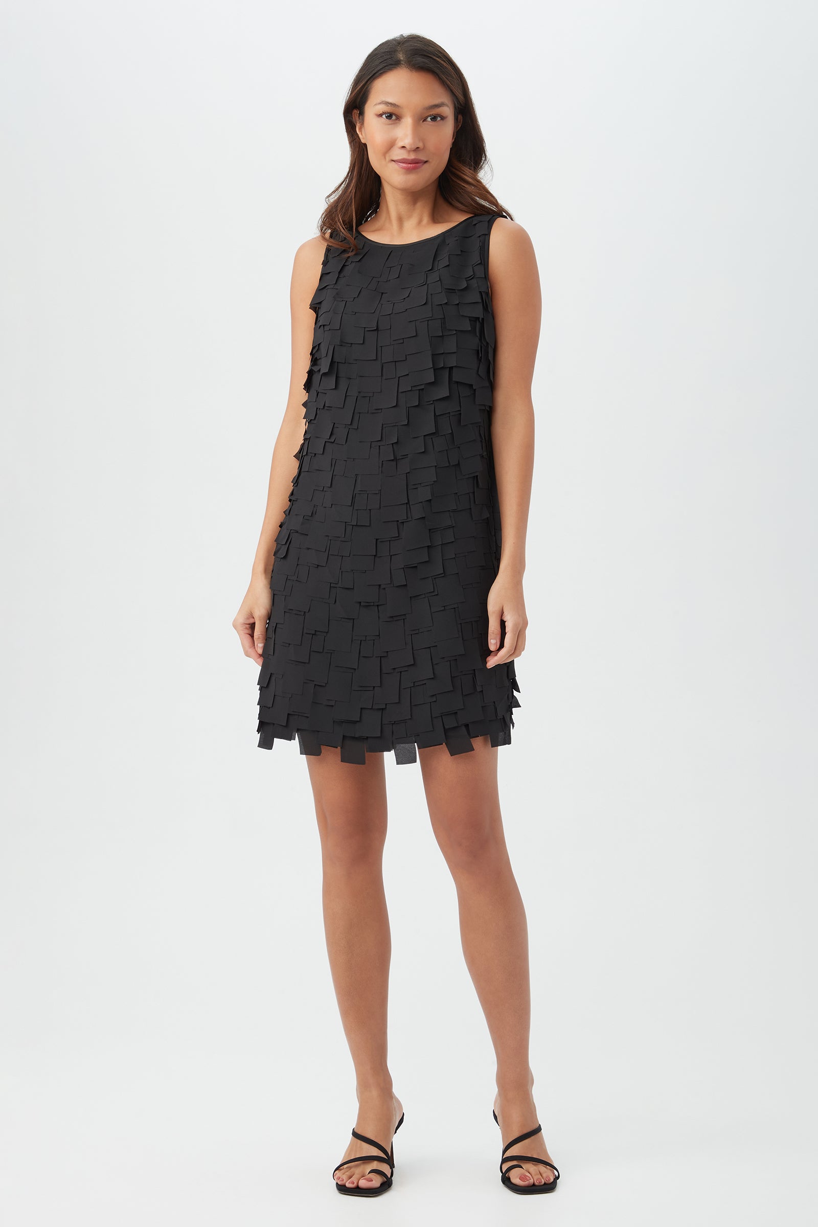 A woman stands against a plain white background wearing the Valdis Dress in black, which features a sleeveless design and a textured, layered square pattern. She completes the look with black strappy heels.