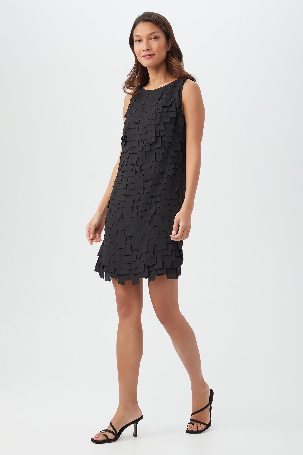 A woman models the Valdis Dress, a sleeveless black dress with a textured square-layered pattern, walking forward in black heeled sandals on a white background and looking at the camera.