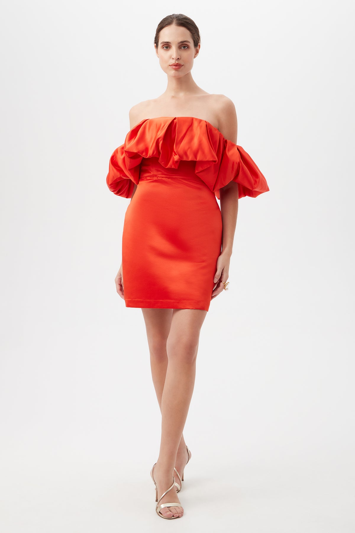 A woman stands against a white background wearing the Nia Satin Off The Shoulder Short Cocktail Dress in Firefinch, featuring voluminous ruffled sleeves. Her hair is pulled back, and she pairs the look with strappy heeled sandals.