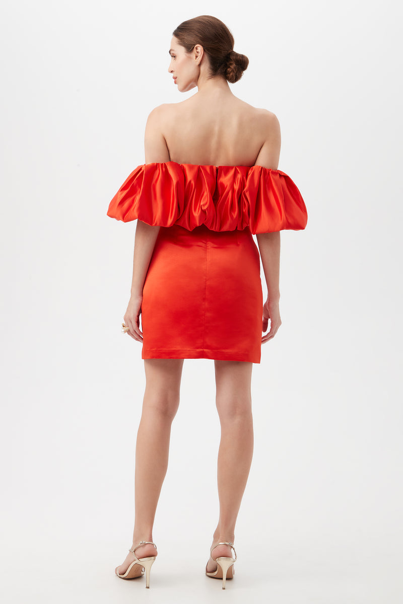 A woman faces away in the Nia Satin Off The Shoulder Short Cocktail Dress, featuring puffed sleeves and an open back. Her hair is styled in a bun, and she wears beige high-heeled sandals against a plain white background.