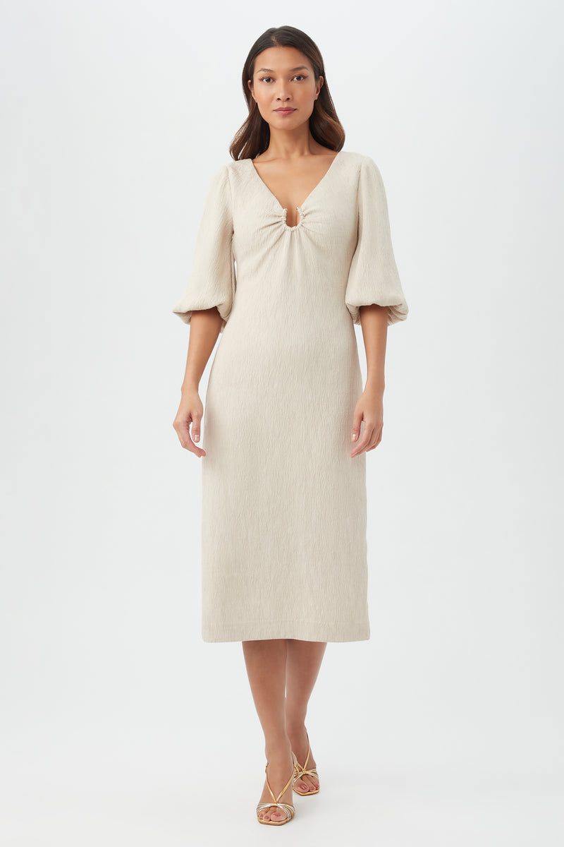 A woman wearing the Madeline Dress in Sahara Stoneâ€â€knee-length with half puff sleeves and a gathered keyhole at the chestâ€â€pairs it with strappy sandals against a plain white background.