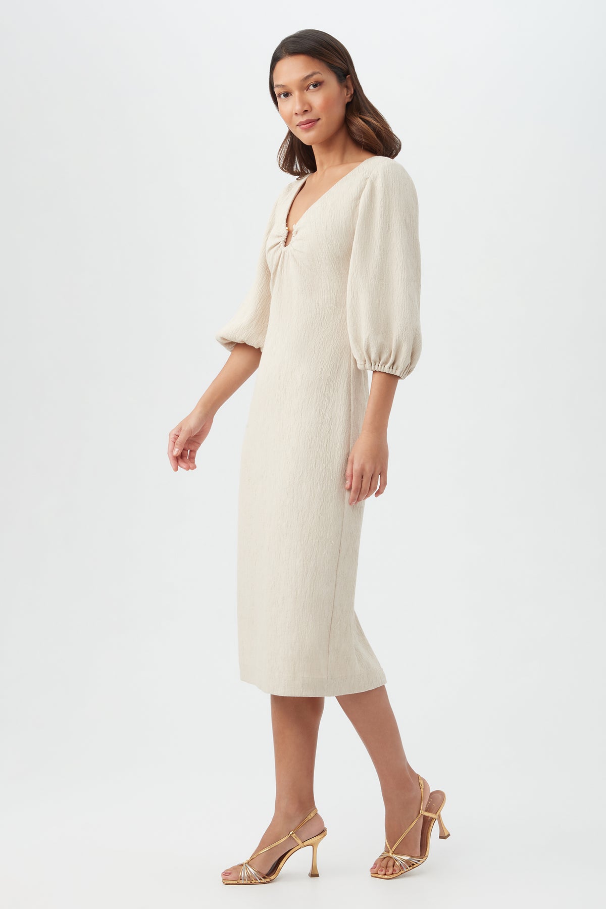 A woman wears the Madeline Dress, a cream-colored, midi-length style with puffed three-quarter sleeves, paired with gold strappy heels, standing against a plain white background.