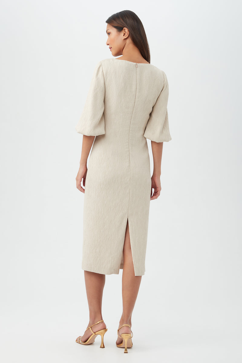 A woman stands against a plain background wearing the Madeline Dressâ€â€a light beige, knee-length dress with three-quarter sleeves and a back slitâ€â€paired with light-colored heeled sandals. She faces away, looking to the side.