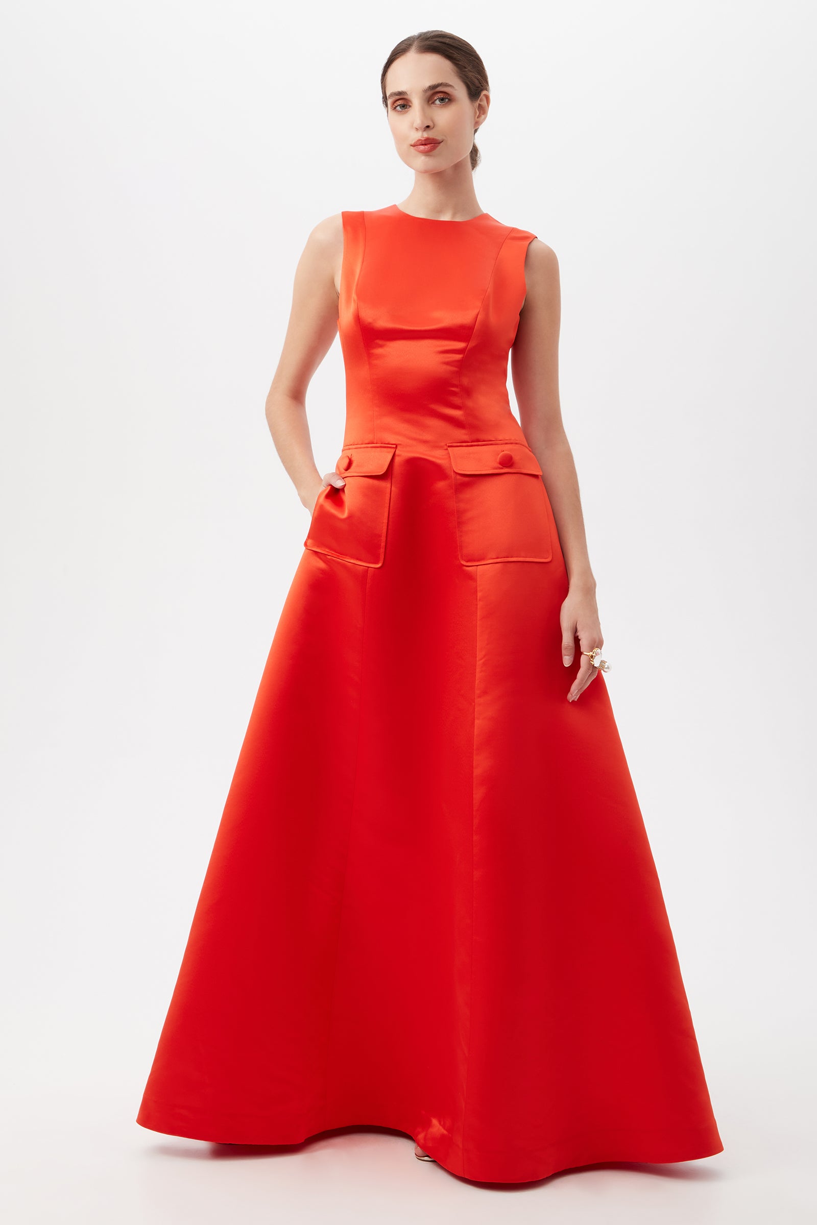 A woman stands against a white background wearing the Guinevere Dress in Firefinch—a sleeveless, floor-length satin gown with a fitted bodice, two front flap pockets, and an A-line skirt. Her hair is pulled back and she wears a ring.